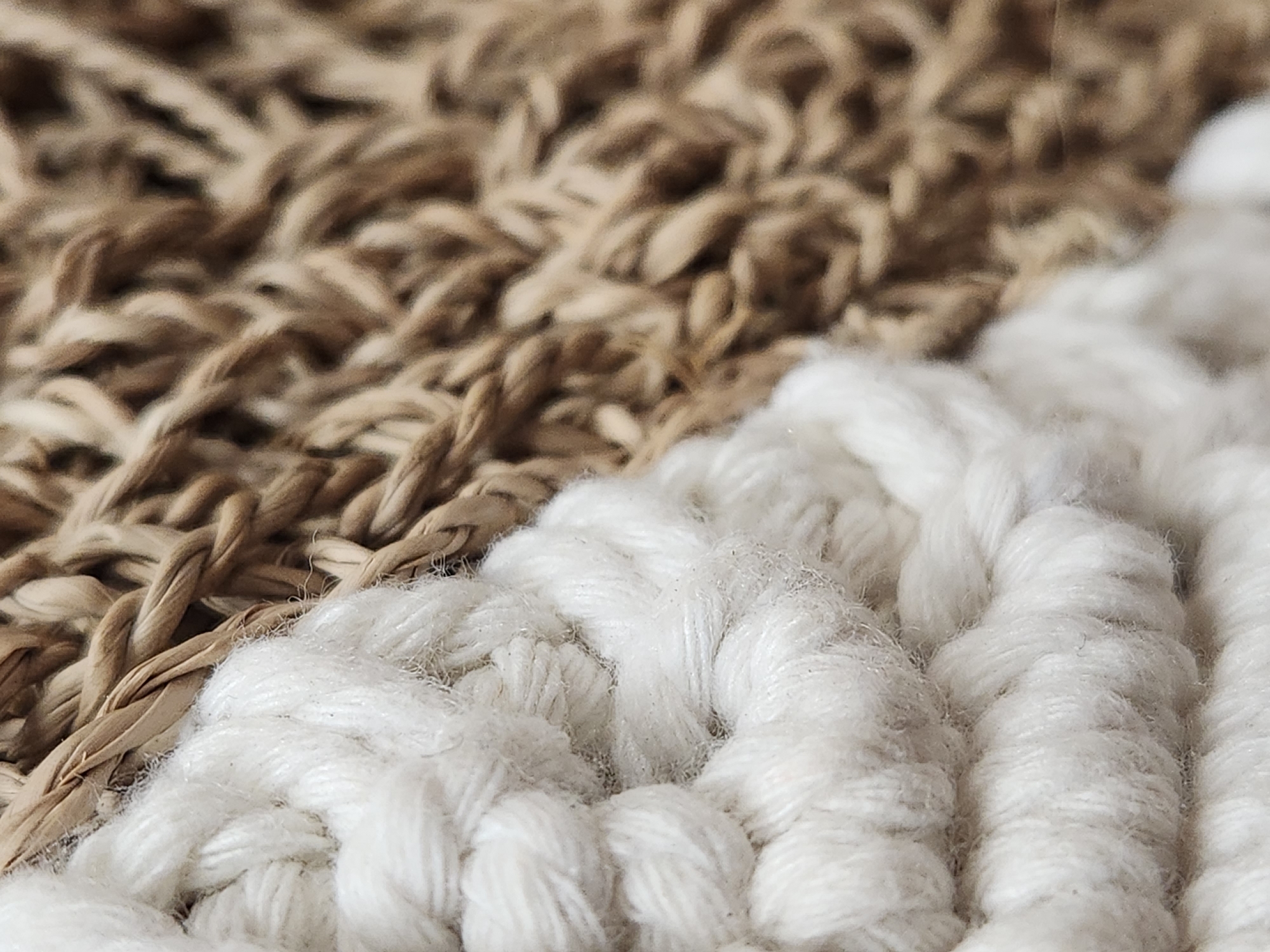 The image shows a close-up, macro photograph of two different woven textures. On the left, a thick, soft, off-white braided rope or cord dominates the frame. The texture is made of multiple strands of yarn twisted together, creating a soft, plush appearance with visible fibers. On the right, a contrasting woven texture of natural, straw-like material is visible. This material is a light brown color and appears to be twisted into thinner strands than the off-white rope, forming a tighter, more rigid weave. The focus is sharp on the textures, highlighting the intricate details of the weaving patterns. There are no people or animals present in the image. The lighting is soft and even, creating subtle shadows that emphasize the depth and texture of the materials. The background is entirely composed of these woven elements, creating an abstract composition focused on tactile qualities. There is no discernible location in Maitland, United States of America, as the image is a tight crop of textured materials.