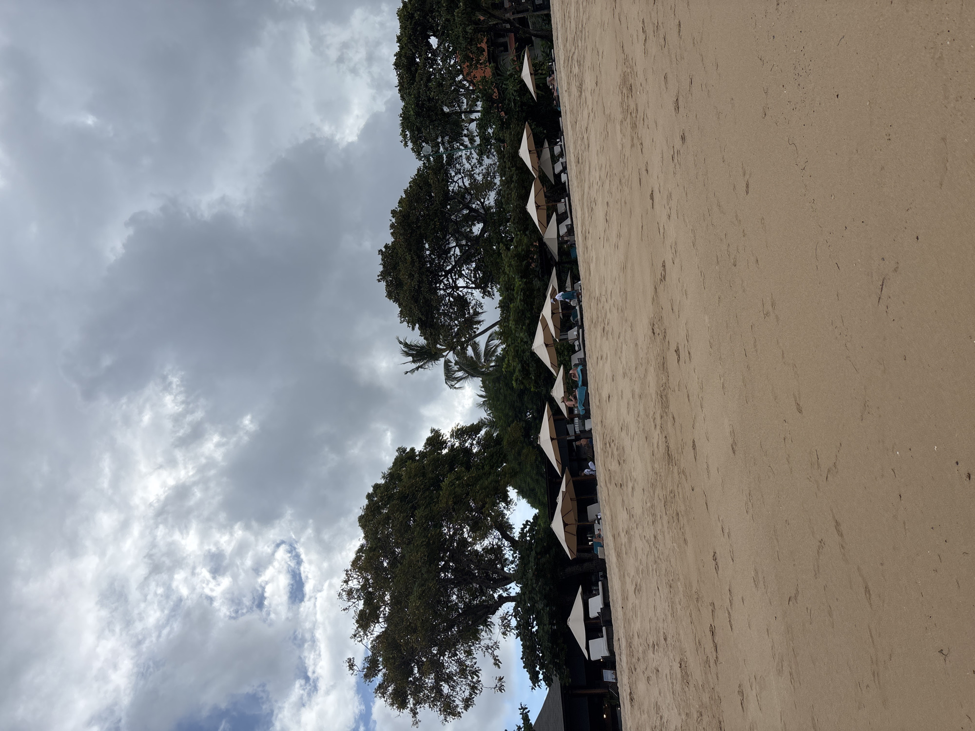 The image depicts a beach scene in Kuta Selatan, Indonesia. The foreground is dominated by a stretch of sandy beach. Along the edge of the beach, there is a line of white umbrellas, presumably shading lounge chairs. Some people are visible under the umbrellas. Behind the umbrellas are various trees. The sky is overcast with thick, gray clouds.