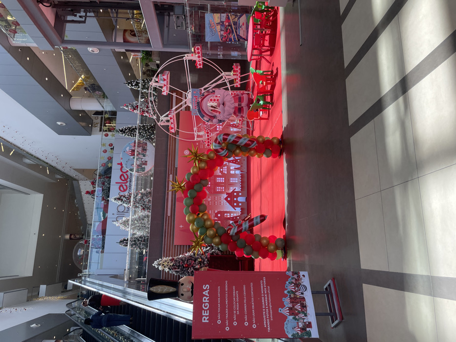 This media file captures an indoor shopping mall, likely located in Luanda, Angola, decorated for a festive occasion. The image is taken from an elevated perspective, looking down at the ground floor where a large Christmas-themed display is set up on a bright red carpet.
The central part of the display features a decorative white Ferris wheel with a Santa Claus cutout in front. A large arch made of red, green, and gold balloons, adorned with candy cane motifs and golden starbursts, frames a background featuring white city silhouette cutouts. Several Christmas trees, some with white "snow" flocking, are positioned around the display. Small red and green tables and chairs are also arranged on the red carpet.
On the left side of the frame, a large nutcracker figure stands next to an escalator. A few people are visible on the escalator, moving between floors. The setting suggests a daytime scene, with natural light entering from above, casting distinct diagonal shadows across the tiled floor on the right side of the image.
A prominent red sign titled "REGRAS" (Rules) lists several regulations in Portuguese: "NÃO TOCAR NOS ARTIGOS" (DO NOT TOUCH THE ARTICLES), "NÃO TRAZER ALIMENTOS E BEBIDAS" (DO NOT BRING FOOD AND DRINKS), "NÃO SE COLOCAR DEBAIXO DA RODA GIGANTE" (DO NOT GO UNDER THE FERRIS WHEEL), "NÃO CORRER PELO CENARIO" (DO NOT RUN AROUND THE SCENE), "NÃO USAR OBJETOS CORTANTES" (DO NOT USE SHARP OBJECTS). It concludes by encouraging visitors to "APROVEITE E GUARDE O MOMENTO TIRANDO FOTOS E MARQUE NOS STORIES @shoppingfortaleza.oficial" (ENJOY AND SAVE THE MOMENT TAKING PHOTOS AND TAG US IN STORIES @shoppingfortaleza.oficial). The overall atmosphere is festive and inviting, designed for family enjoyment and photo opportunities.