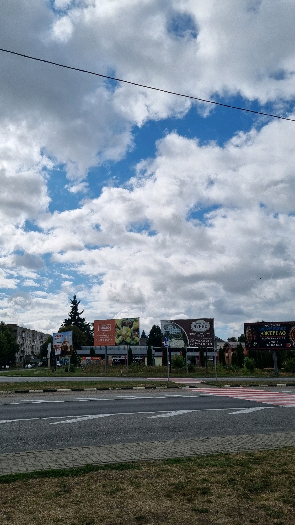 This is a vertical, eye-level, outdoor, daytime, full shot of a street scene in Dubno, Ukraine, with a focus on billboards. The sky is filled with dramatic white and gray clouds against a bright blue background. A thin black wire stretches diagonally across the upper portion of the frame. Below the sky, a line of billboards stands prominently in the middle ground. From left to right, the billboards advertise: "НЕ ЗВАЖАЙТЕ" (Don't be afraid) with a picture of a person on a horse, a "КОМО" (Komo) brand with an image of cheese and fruits, a hotel called "ГОТЕЛЬ ДУБНО" (Hotel Dubno) with a picture of a building and contact information, and a restaurant called "РЕСТОРАЦІЯ ШЕВЧЕНКОВЕ ДЖЕРЕЛО" (Shevchenkove Dzerelo Restaurant) with an image of traditional Ukrainian food and contact information. In the background, a line of apartment buildings and trees is visible. The foreground features a road with white lane markings and a pedestrian crossing, bordered by a patch of dry grass. No people are visible in the frame.