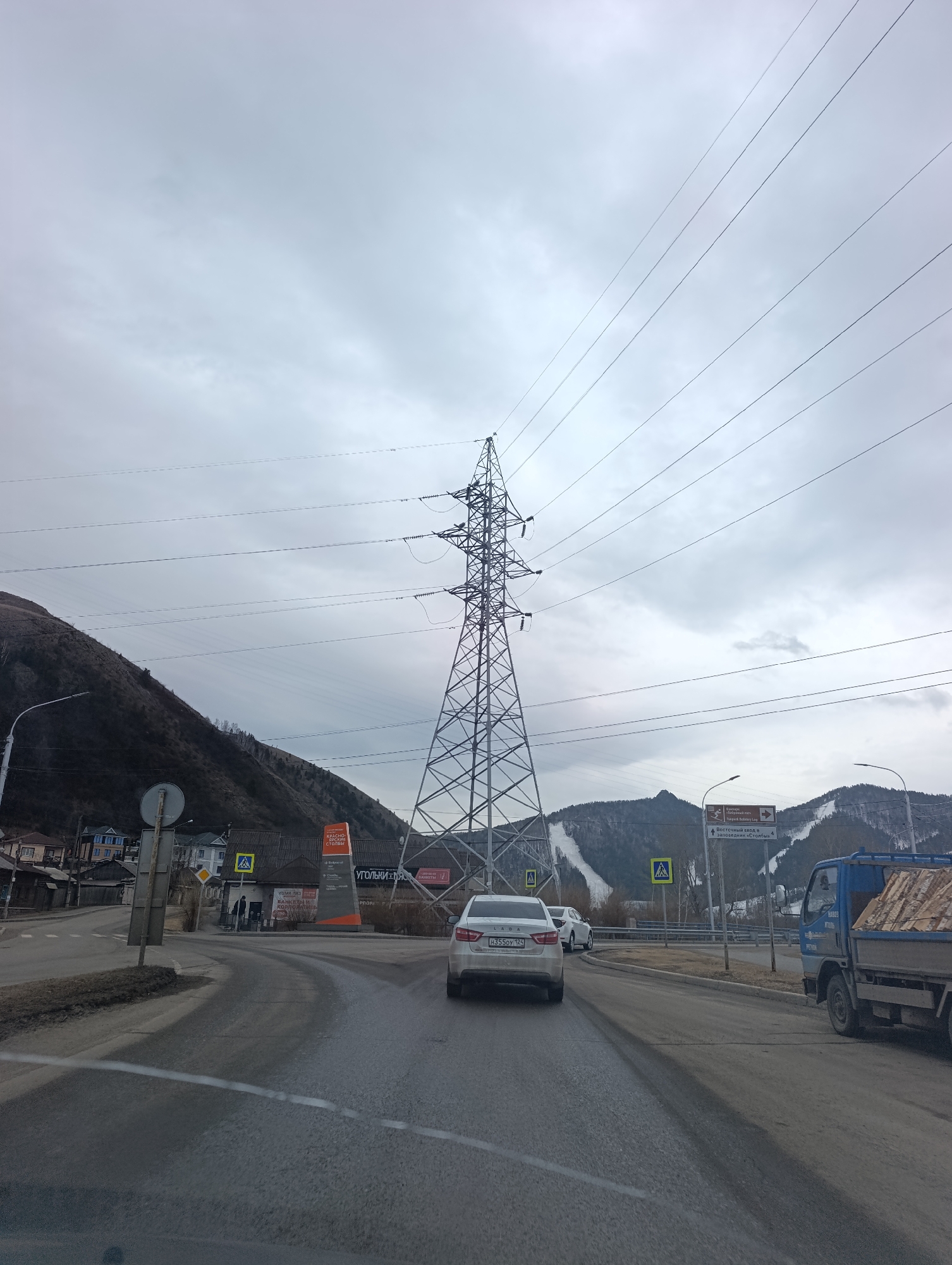 The image captures a street scene in Krasnoyarsk, Russia. An electrical tower stands prominently, intersected by several power lines running across the sky. In the foreground, a road curves, with a white car displaying the license plate "H355OY 124" driving on it. There is also a blue truck on the right side of the road. Street signs are visible, including one indicating directions to the "Funpark Bobrovy Log" and the "Vostochny Entrance to the Stolby Reserve." A building with a sign displaying "Угольки ЛЯС" is on the left. Mountains are present in the background, with ski slopes visible on one of them. The sky is overcast with a layer of grey clouds.