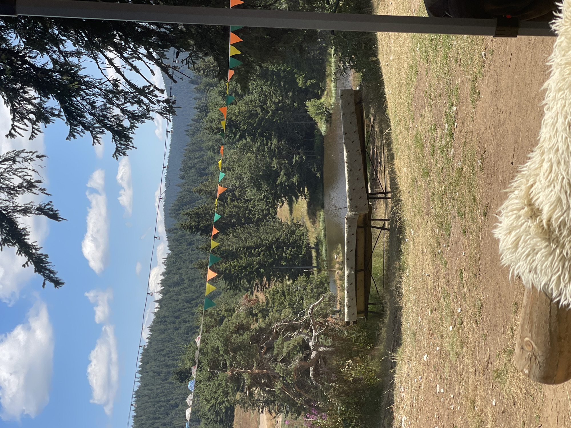 The image depicts a serene outdoor scene in the city of Smolyan, Bulgaria, likely a campsite or a picnic area nestled within a forest. In the foreground, a rustic wooden log is visible, covered with a fluffy white throw. Behind it, two elongated benches with worn, patterned upholstery are set up on a patch of dry grass. Beyond the benches lies a tranquil body of water, a small lake or pond, reflecting the clear blue sky dotted with white, fluffy clouds. The banks of the water are lined with lush green vegetation and mature pine trees. In the middle ground, a string of colorful triangular flags, red, yellow, green, and orange, is stretched across the scene, adding a festive touch. Further in the background, a dense forest covers the rolling hills, with a hazy mountain range in the far distance. The overall atmosphere is peaceful and natural, suggesting a place for relaxation and enjoying the outdoors.