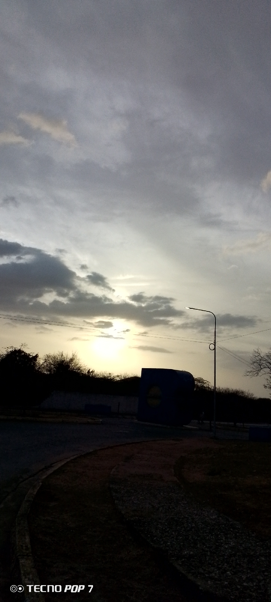 A landscape photo taken at dusk or dawn, depicting a scene in or near a city. The sky is filled with grey and white clouds, with a hint of sunlight peeking through. In the middle ground, there is a dark silhouette of trees or bushes and a large blue object, possibly a container or billboard. A street light is also visible. The foreground consists of a path or road with gravel or dirt on the edges. The location is Altagracia, Venezuela. The camera model used is a Tecno Pop 7. There are no people visible in the photo.