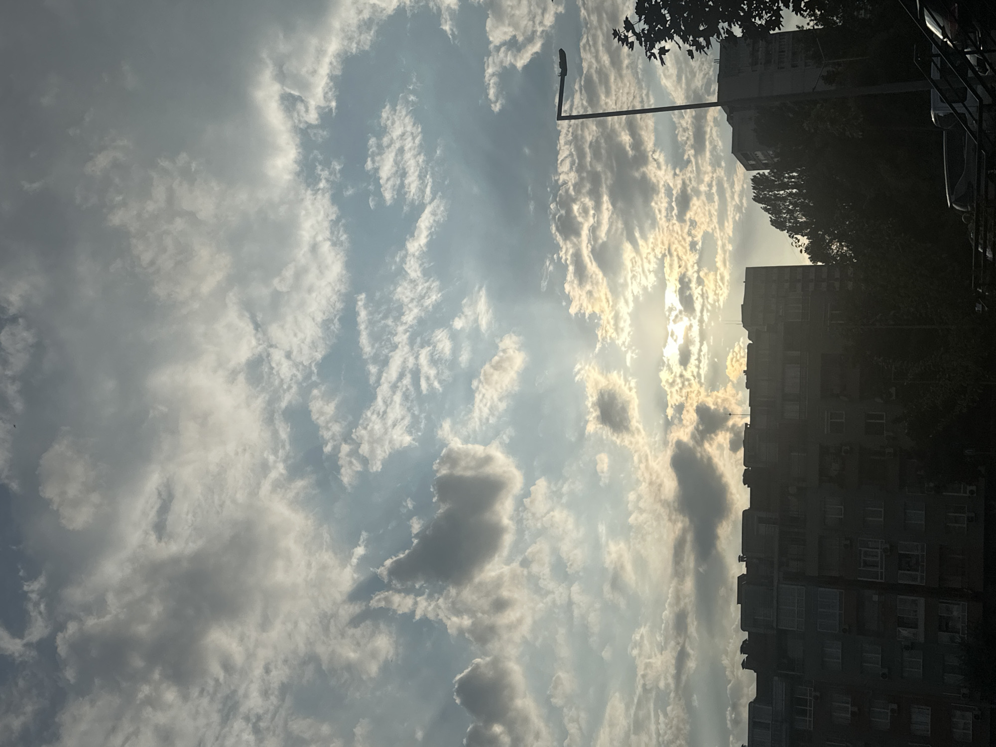 The image shows a view of buildings against a cloudy sky with the sun partially visible behind the clouds. A streetlight is visible on the right side of the image. There are trees in the foreground and midground, and cars are parked in the lower right corner. The buildings appear to be apartment blocks, with windows and balconies visible. The sky is filled with a mix of gray and white clouds, with patches of blue sky visible. The sun is casting a warm glow through the clouds, creating a dramatic effect. The overall impression is one of an urban environment on a partly cloudy day.