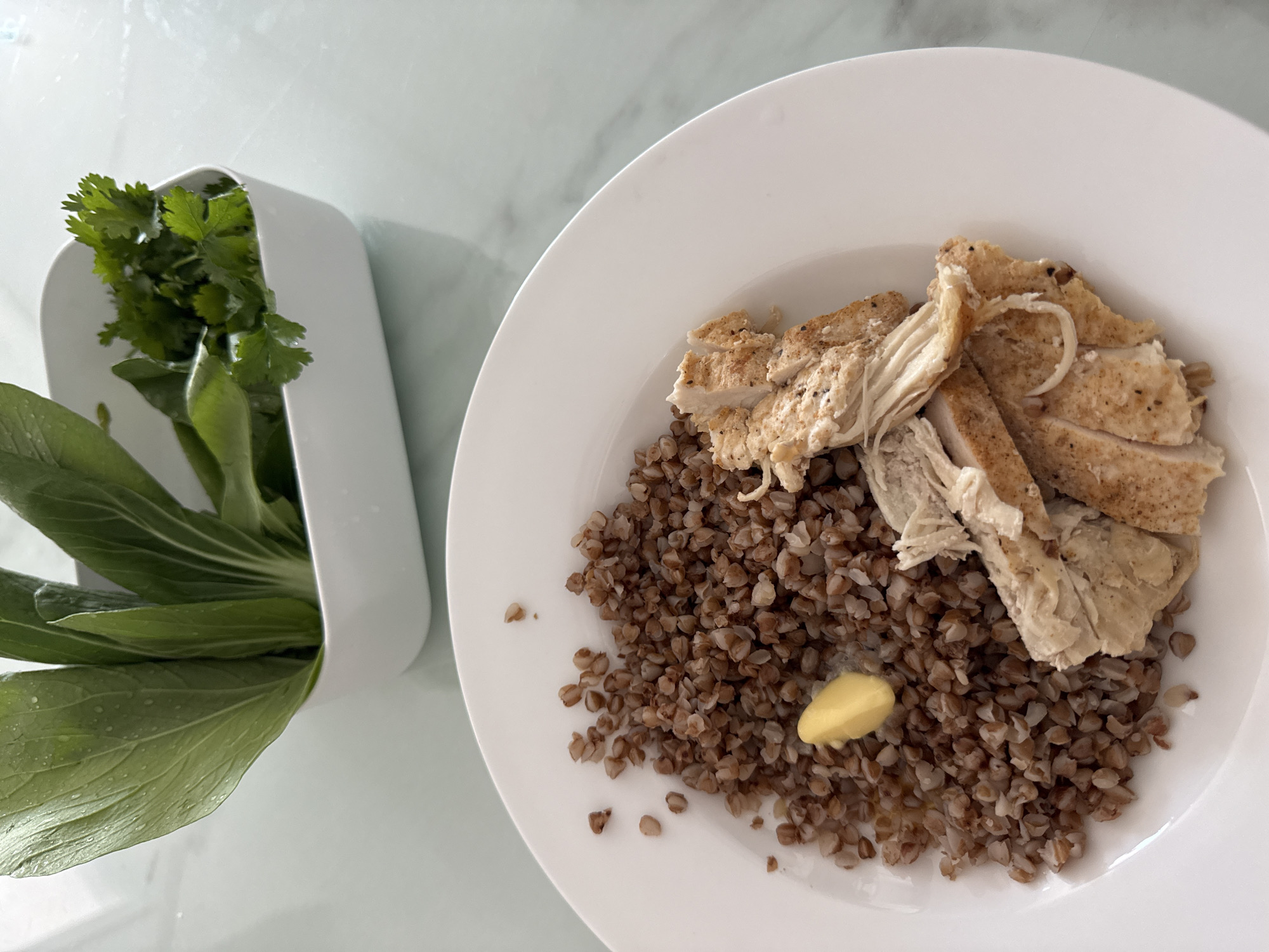 On a light-colored surface, a white plate containing buckwheat, sliced chicken, and a pat of butter is positioned next to a white container holding bok choy and cilantro. The image suggests a meal setting. There is no evidence to confirm that this image was taken in the city of Bac Nha Trang Ward, Vietnam.