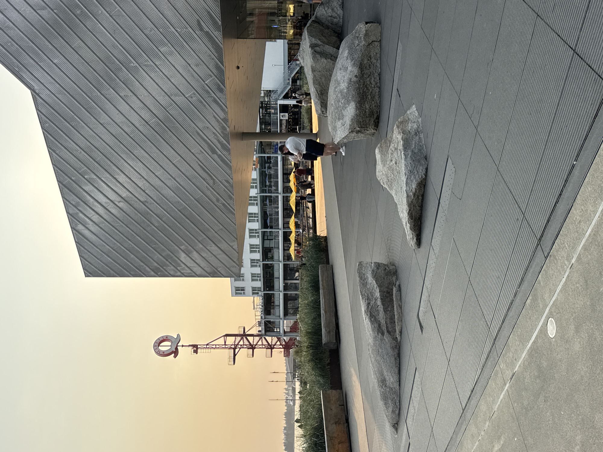This image depicts a view of the waterfront in the city of North Vancouver, Canada, likely at dusk or during a hazy sunset, given the warm, diffused light. In the foreground, several large, rough-hewn grey rocks are placed on a dark grey tiled plaza with a grid pattern. To the left, a wooden planter box filled with tall, dry grass separates the plaza from a body of water in the distance. A red, lattice-like structure with a prominent red "Q" sign on top stands tall against the sky. Behind this structure, a multi-story white building with visible windows and balconies lines the waterfront. Yellow umbrellas are visible on outdoor seating areas in front of this building.
The middle ground shows a few individuals walking and standing on the plaza and near the building. Two people are facing away from the camera, one in a white polo shirt and dark shorts, the other in dark clothing. The background features a body of water with a boat or ferry visible on the horizon, and a tree line on the far shore. The sky is a pale orange-yellow, suggesting atmospheric haze. The architecture of the building in the foreground is modern, with a metal facade featuring diagonal lines.