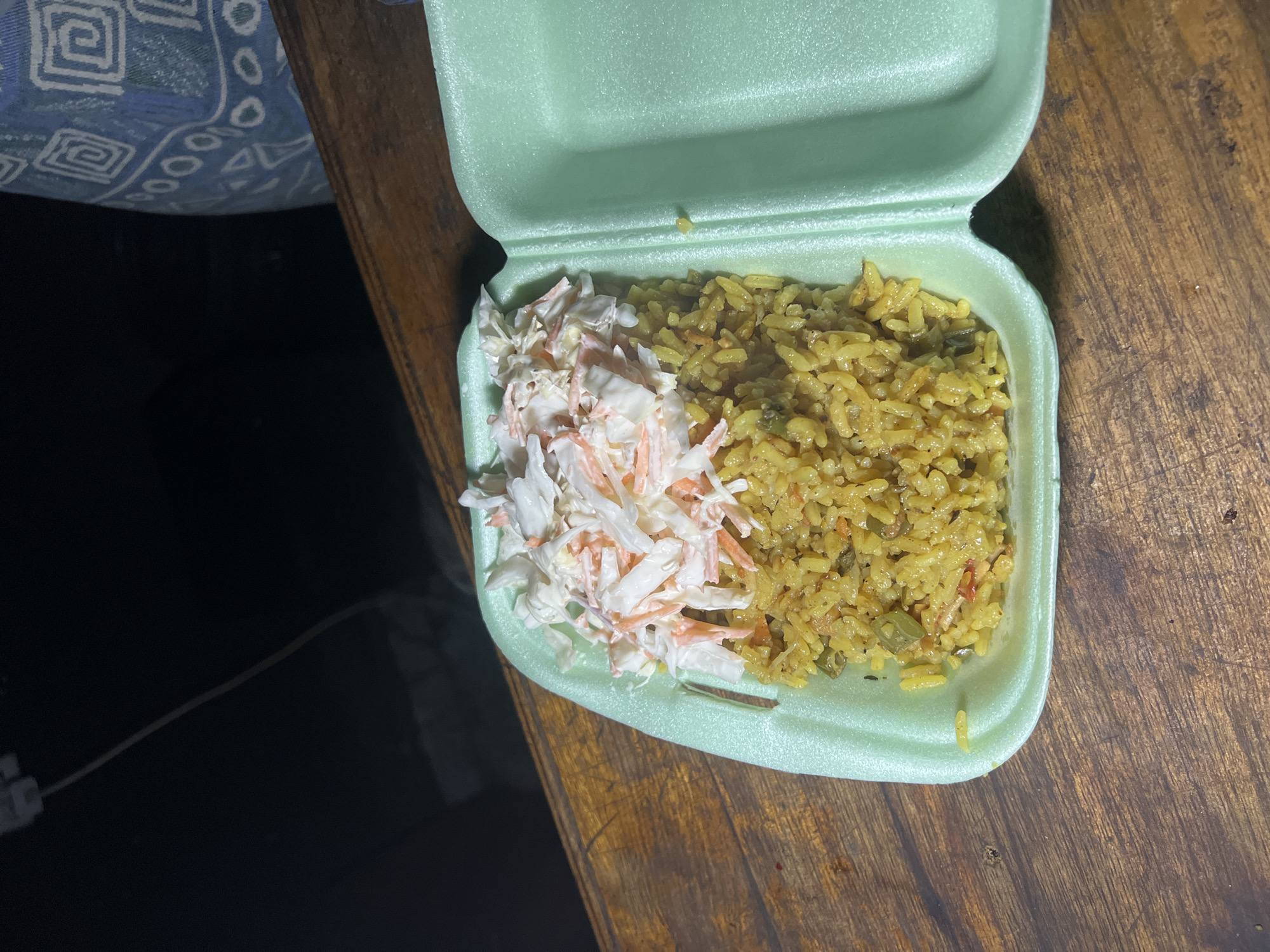 A takeaway container of food is on a wooden surface. The container is pale green and appears to be made of styrofoam. Inside, the container is divided into two sections. The left section is filled with a portion of yellow rice mixed with visible chopped vegetables, possibly carrots and green beans. To the right of the rice is a pile of coleslaw, consisting of shredded cabbage and carrots with a creamy dressing. The background is dark and out of focus, but a portion of a blue and white patterned fabric is visible on the right. The wooden surface is worn and appears to be a table or counter. The scene is dimly lit, suggesting it may be nighttime or an indoor setting with low light. There are no people visible in the image. The location is specified as Durumi, Nigeria.