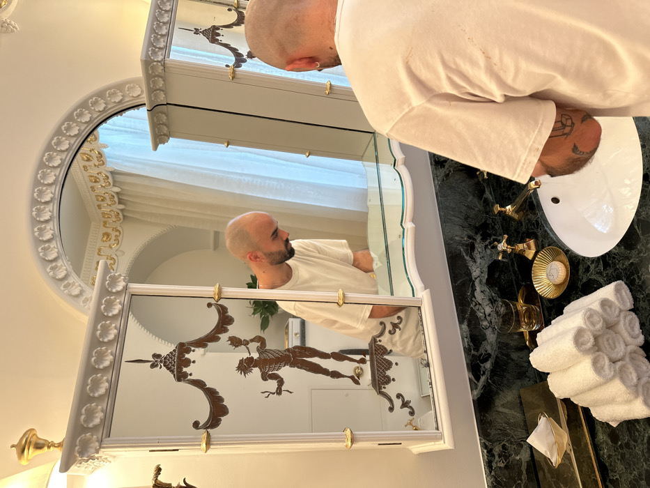 The image depicts a bathroom scene in the City of London, United Kingdom. A bald male subject with a beard, wearing a white t-shirt, is observed from behind, leaning forward over a white oval sink. His reflection is visible in the central mirror, showing his head turned downwards towards the basin. Tattoos are visible on the subject's right forearm, including a peace sign graphic.
The sink basin is set into a dark green marble countertop with white veining. Gold-colored, traditional cross-handle faucets are installed. To the left of the sink, on the countertop, are a stack of white rolled towels, a gold-colored tray holding a tumbler with an amber liquid, a gold shell-shaped soap dish containing a white bar of soap, and a gold tissue box holder with white tissues.
Above the countertop is an ornate, white-framed mirror unit comprising a central mirror and two mirrored cabinet doors. This unit is detailed with gold accents and decorative white molding featuring leaf motifs. The left cabinet door features an etched or painted brown motif of a standing skeletal figure holding an object, positioned beneath an elaborate canopy. Architectural molding with gold leaf elements is present above the mirror unit, and a white curtain is visible in the background reflection.