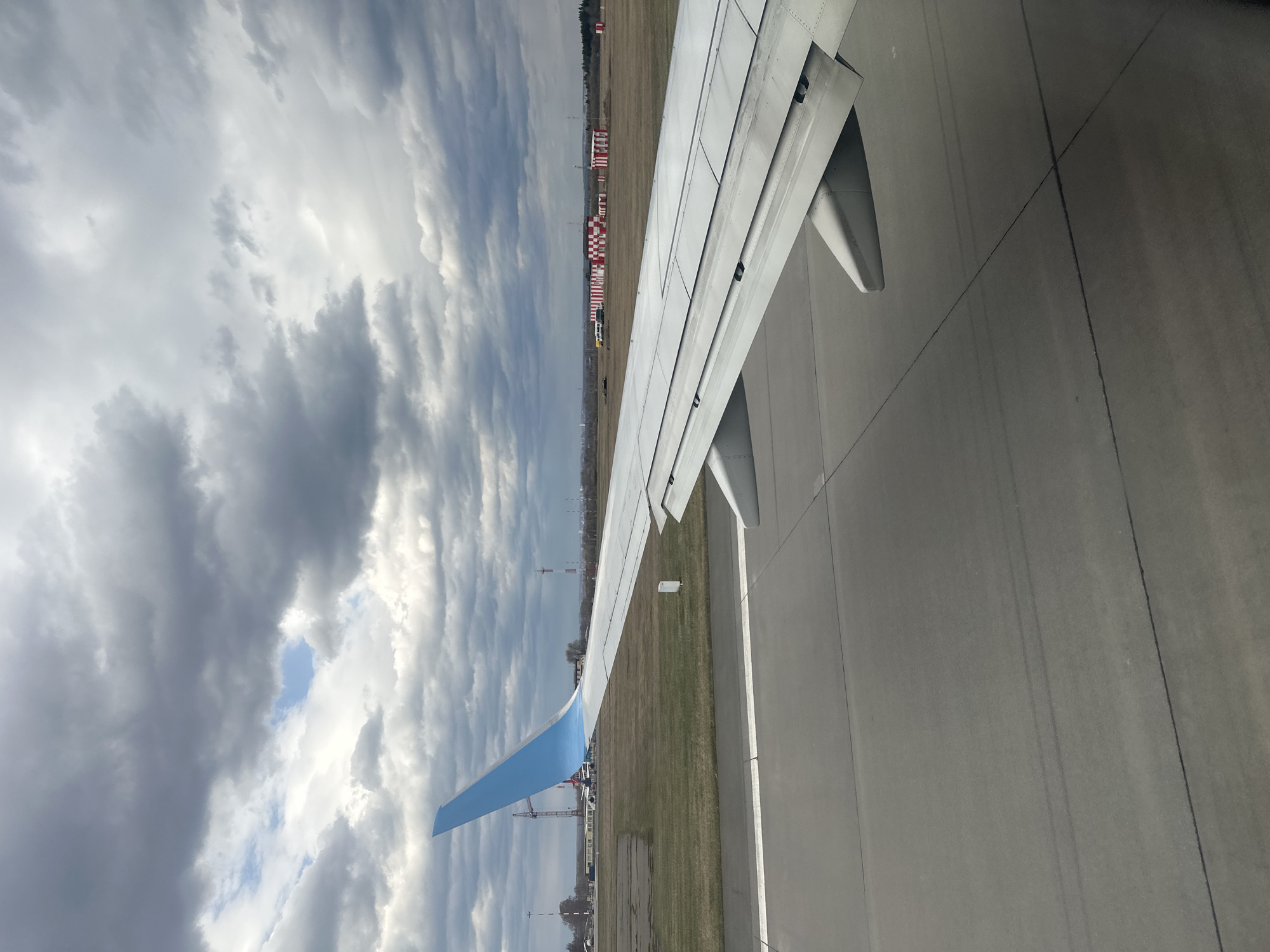 This media file displays a view from an airplane window, showing an aircraft wing on the ground at an airport. The image is oriented vertically.
On the left side of the frame, a cloudy sky dominates, featuring dense grey and white clouds with visible patches of brighter blue sky breaking through. The airplane's wing, predominantly white with a light blue wingtip, extends diagonally across the middle of the image, from the lower-left towards the upper-right. The wing shows mechanical details and panels.
To the right of the wing, a paved surface, likely an airport runway or taxiway, is visible, marked with subtle lines. Below the wing and beyond the paved area, a strip of dry, light brown grass separates the foreground from the background elements. In the distance, various airport structures can be seen, including distinctive red and white checkered markers, some smaller buildings, and a line of trees under the overcast sky.
The scene depicts an airplane on the ground, possibly taxiing or waiting for takeoff/landing. The time of day appears to be daytime, with an overcast sky. There is no discernible text visible in the image. The location is contextualized as Bolshoye Savino, Russia.