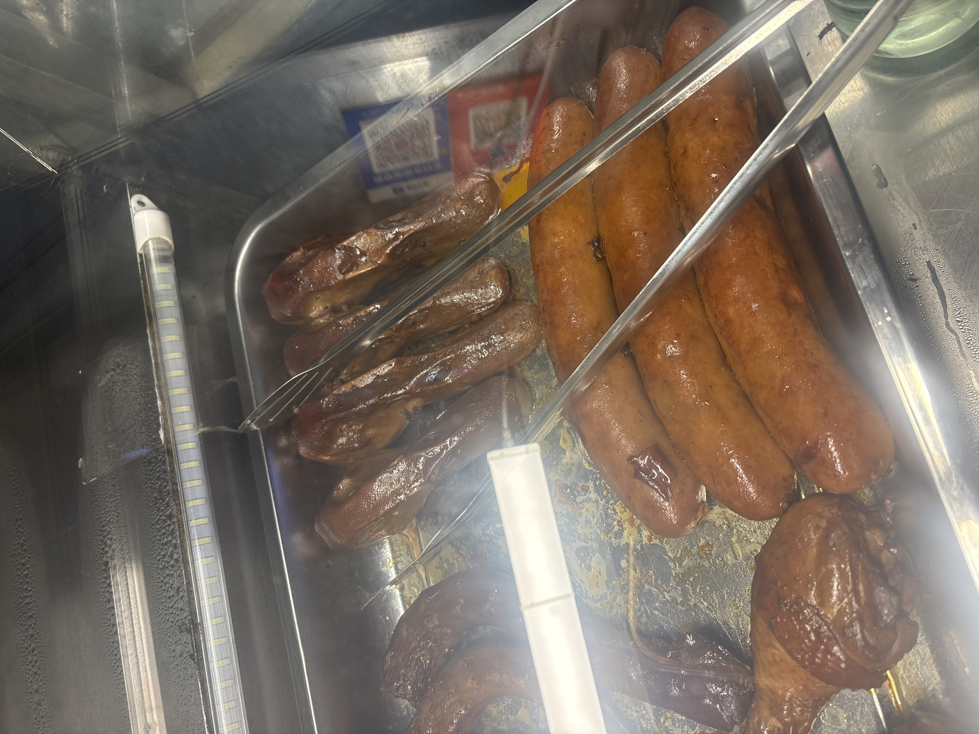A metal tray filled with cooked meats, including what appears to be duck necks and sausages, sits inside a refrigerated display case. The tray has some grease and condensation. A pair of metal tongs rests on top of some of the sausages. The display case is lit by a fluorescent strip light. In the background, a small red and white package with Chinese characters is visible. The overall environment appears to be a food service establishment, possibly a deli or market stall, located in the Suzhou Industrial Park, China.