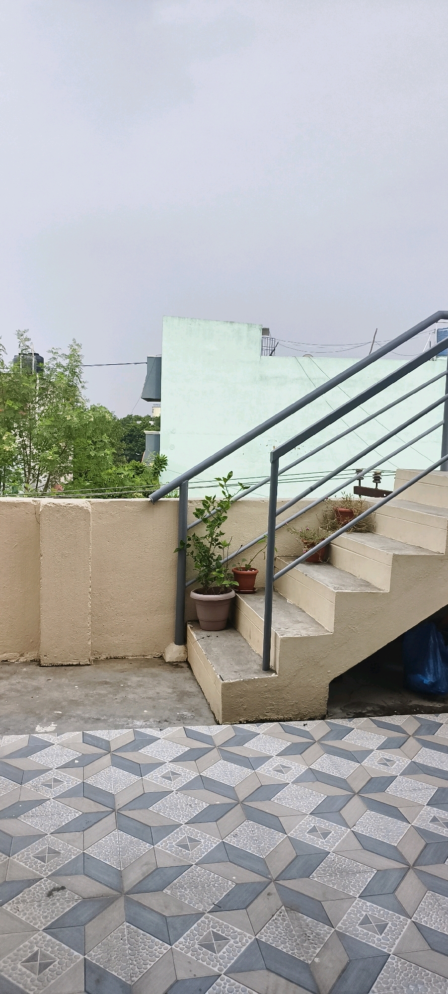 The image shows an exterior view of a rooftop or balcony area in Kundrathur, India. In the foreground, there is a patterned tile floor with a geometric design of interlocking grey, white, and blue diamond shapes, some with a pebble-like texture. To the right, a concrete staircase with a light beige painted railing leads upwards. The staircase has several steps, and the risers are also painted beige. A metal railing with multiple parallel bars runs along the side of the stairs. On the stairs, there are two potted plants. The larger pot contains a green leafy plant, and a smaller pot with dry plants is situated above it.
To the left of the staircase is a light beige painted concrete wall, which appears to be a boundary or retaining wall. Behind this wall, there are trees with lush green foliage and a water tank on a building in the background. Further back, a light green painted building with some architectural features is visible, along with electrical wires and a metal structure on its roof. The sky is overcast and appears a pale grey, suggesting a cloudy day. The overall scene depicts an outdoor urban environment, likely part of a residential building.