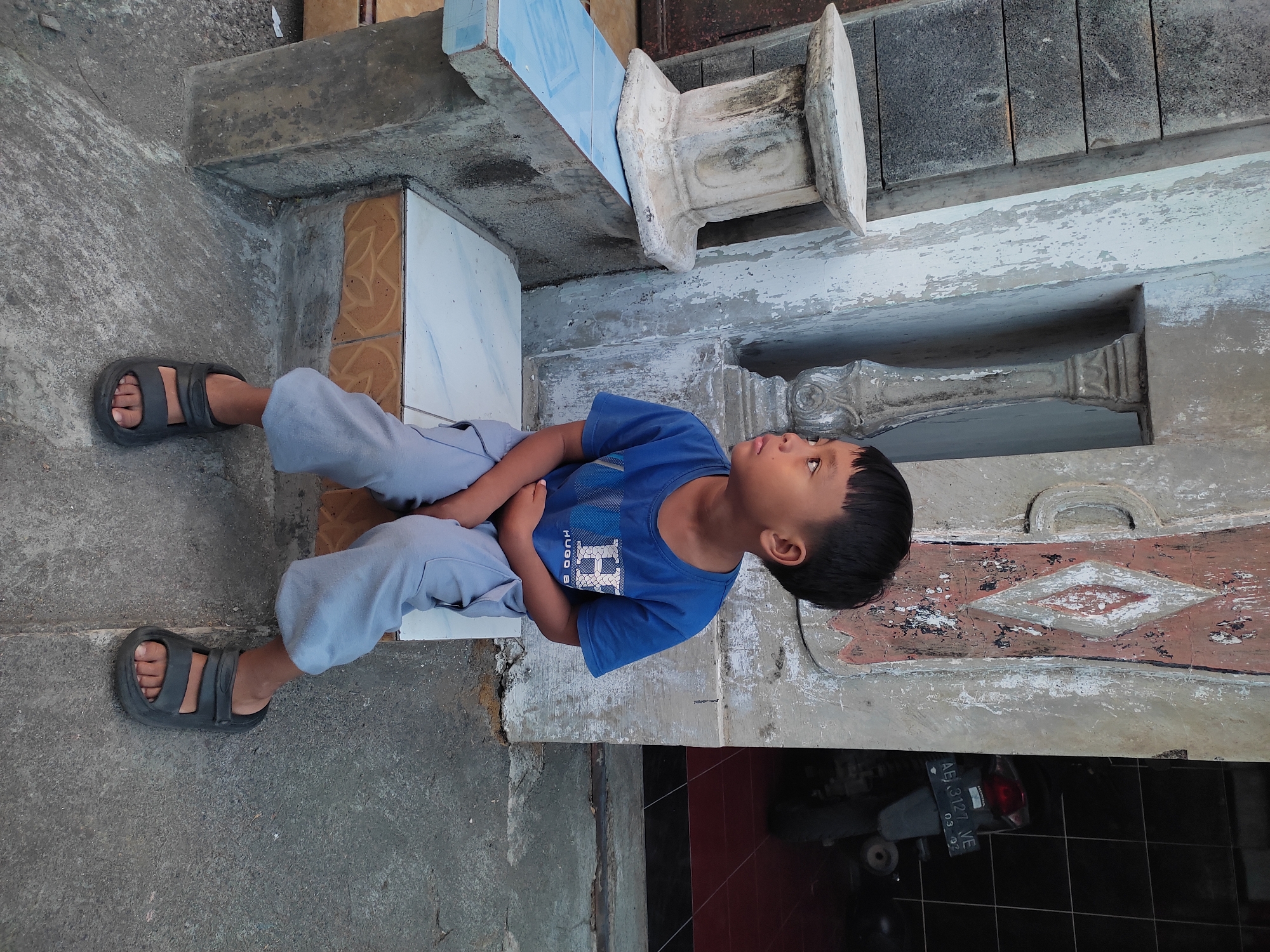 A young boy with short black hair sits on a concrete step in front of a building. He is wearing a blue t-shirt with a white "H" and "HUGO BOSS" logo, and light gray pants. He has black sandals on. The building behind him appears to be a house or shop, with weathered walls and decorative elements. A motorcycle with a license plate that reads "AE 3127 VE 03-26" is visible inside the doorway. The ground is made of concrete and gravel. The scene is set in Ponorogo, Indonesia.