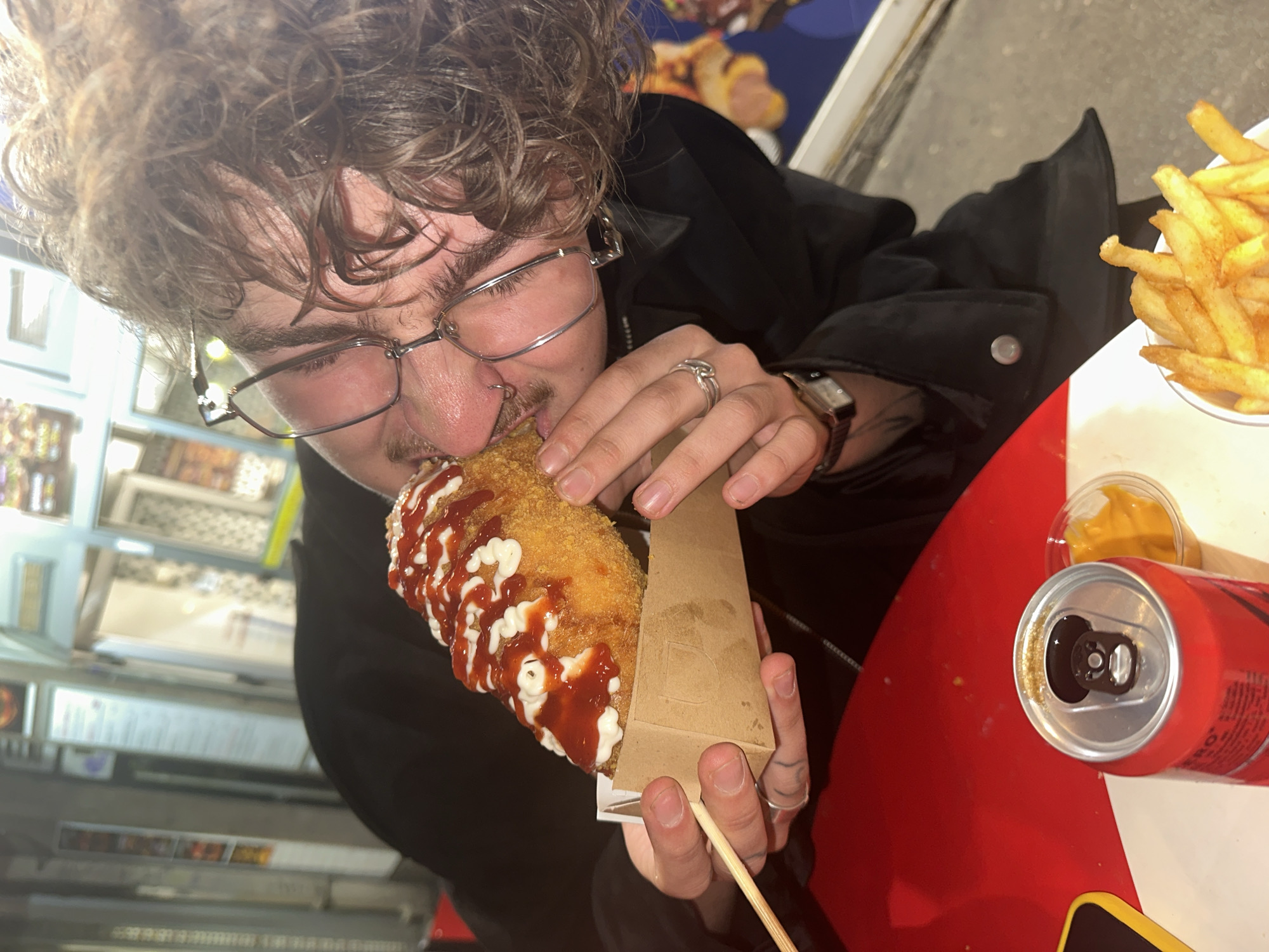 In this close-up, eye-level shot, a person with curly brown hair and glasses is leaning in to eat a corn dog, which is topped with ketchup and mayonnaise. The corn dog is held in a brown paper tray by the person's hands. The person is wearing a black jacket, a watch on their left wrist, and rings on their fingers. In the foreground, a red can and a small container of yellow sauce are visible on a red surface. The background suggests an outdoor setting with lights and some structures resembling food stalls, possibly in Belgrade, Serbia.