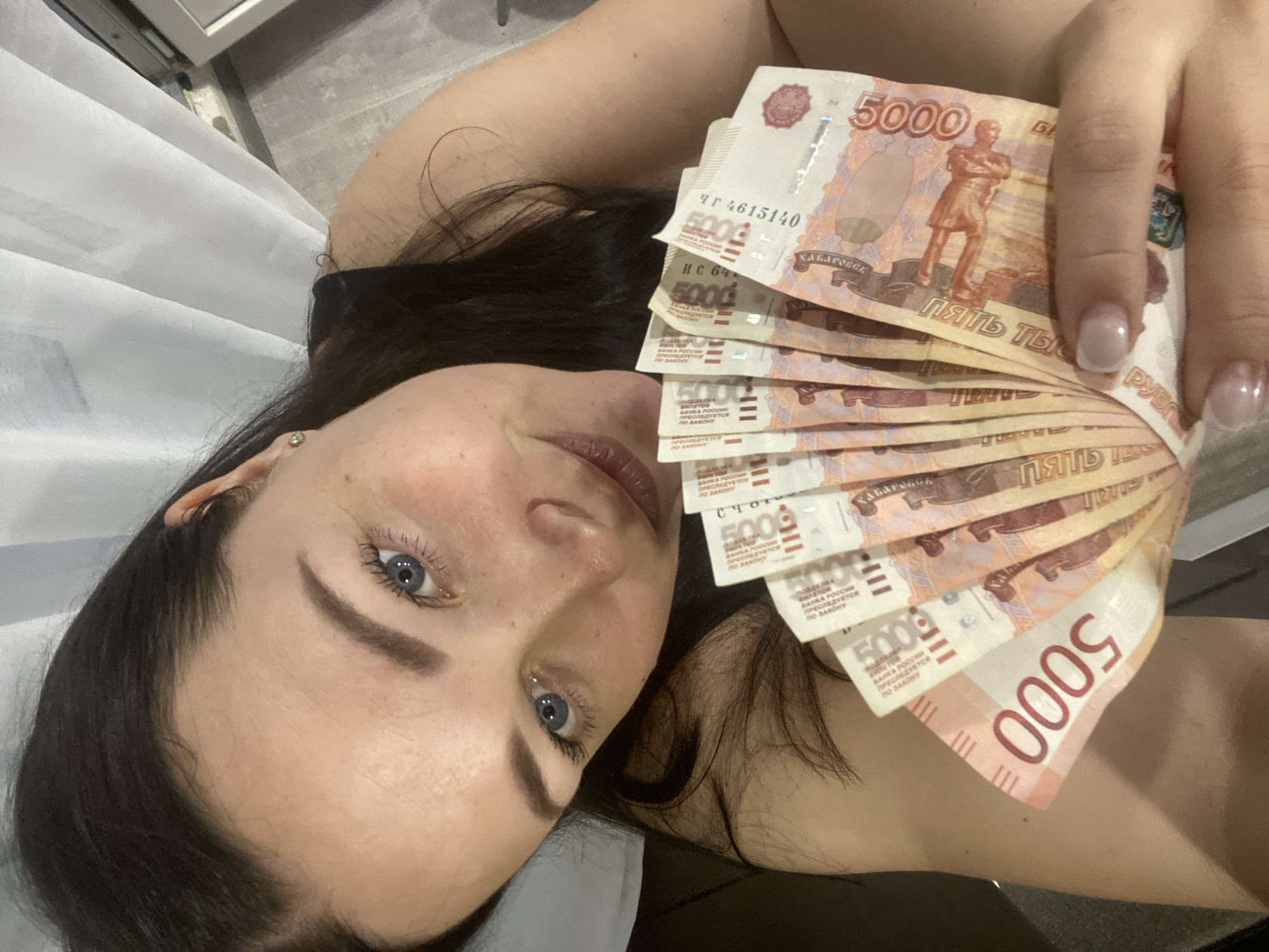 A young woman is depicted in a close-up, top-down shot, appearing to lie down indoors, likely in Sochi, Russia. She has long dark hair, light blue eyes, and well-defined eyebrows, looking directly at the camera with a neutral to slightly smiling expression. A small stud earring is visible in her left ear. Her right hand, adorned with manicured nails featuring a light polish, holds a fan of several 5000 Russian Ruble banknotes. The background includes a sheer white curtain or fabric on the left and a light-colored surface, possibly flooring or a wall, to the right, suggesting an indoor setting.
The primary activity is the woman displaying the banknotes. Visible text on the 5000 Ruble notes includes the denomination "5000," "ПЯТЬ ТЫСЯЧ РУБЛЕЙ" (Five Thousand Rubles), "БАНКА РОССИИ" (Bank of Russia), "ПРЕСЛЕДУЕТСЯ ПО ЗАКОНУ" (Punishable by Law), and "ХАБАРОВСК" (Khabarovsk). Partial serial numbers such as "ЧГ 4615140" and "ИС 64" are also discernible on some of the bills. The time of day is not evident, but the lighting suggests an interior environment.