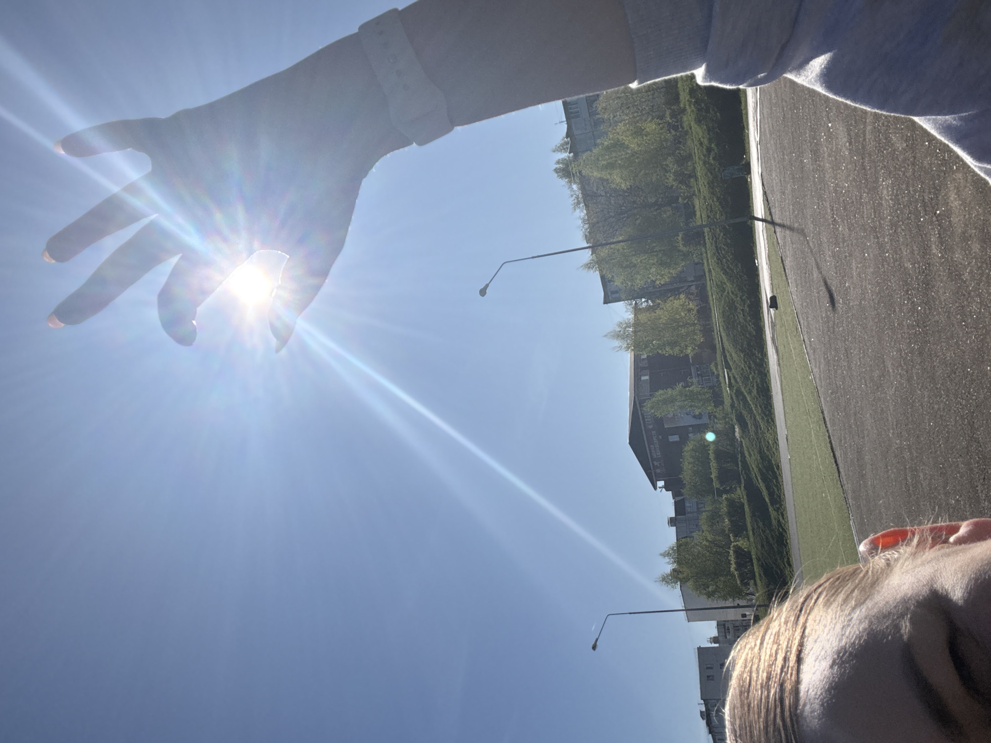 A hand is held towards the sun, obscuring it, against a blue sky. Below the hand, there are buildings, trees and a field or sports track with a fence, along a road with lampposts. This image appears to have been taken in Chaikovsky, Russia. A person's hair and ear is visible.