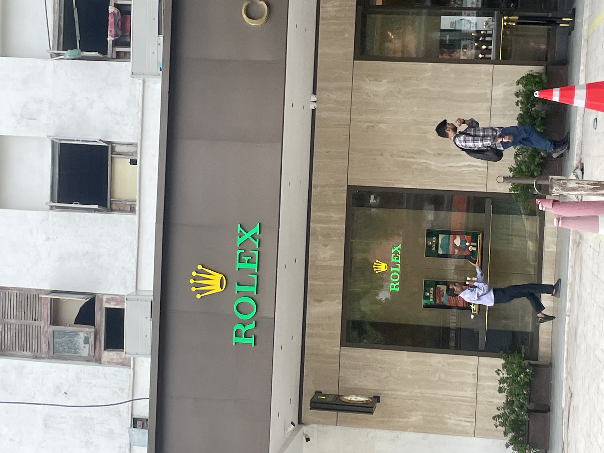 The image shows the exterior of a Rolex store in New Delhi, India. The store has a large, dark gray facade with the prominent green "ROLEX" logo and a gold crown above it. Below the main sign, large glass windows display Rolex watches. Two people are visible: a man in a light blue shirt and dark pants is walking towards the store entrance, holding what appears to be a phone, and a man in a plaid shirt and blue jeans is walking away from the store, carrying a backpack. Outside the store, planters with green foliage and flowers are arranged along the sidewalk. The building itself appears to be multi-story, with some windows on the upper floors appearing weathered and in disrepair, featuring wooden shutters. The overall scene depicts a busy street in a commercial area of New Delhi.