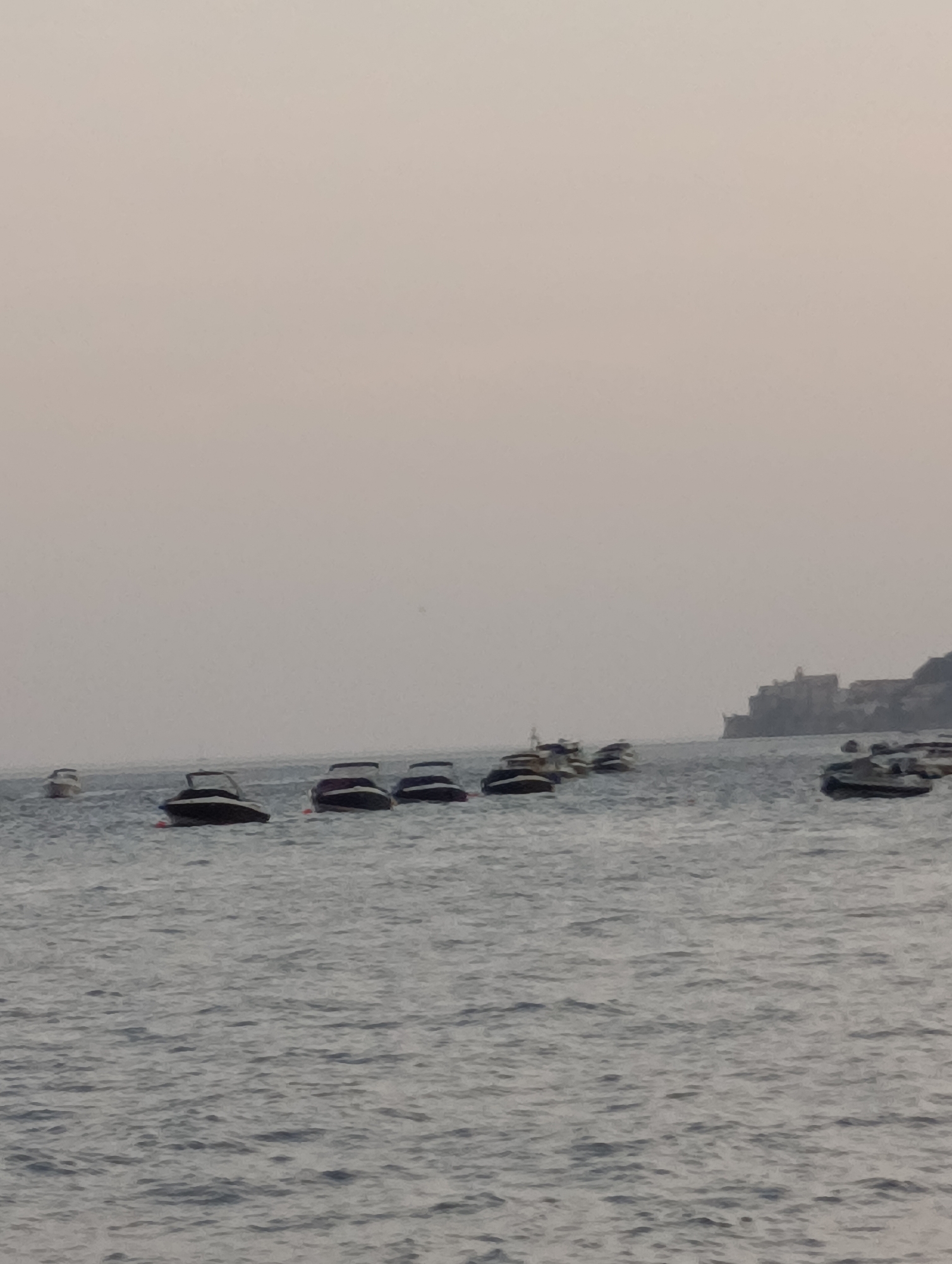 The image shows a bay or harbor area with several small motorboats anchored on the water. The sky is overcast, and the water is slightly choppy. In the distance, a coastline with buildings and cliffs is visible. The overall impression is a calm, perhaps early morning or late afternoon scene. The location is identified as the Bay of Setúbal, Portugal, a well-known port city with a scenic coastline.