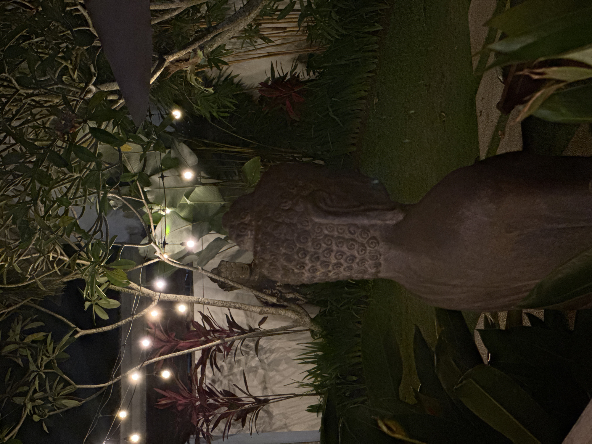 The image depicts an outdoor scene, likely a garden, in Kuta Selatan, Indonesia, at night. A stone statue of a human figure is prominently featured, surrounded by dense foliage, including trees, grass, and broad-leafed plants. String lights are strung among the tree branches. The background shows a wall and possibly a building. No people are visible.