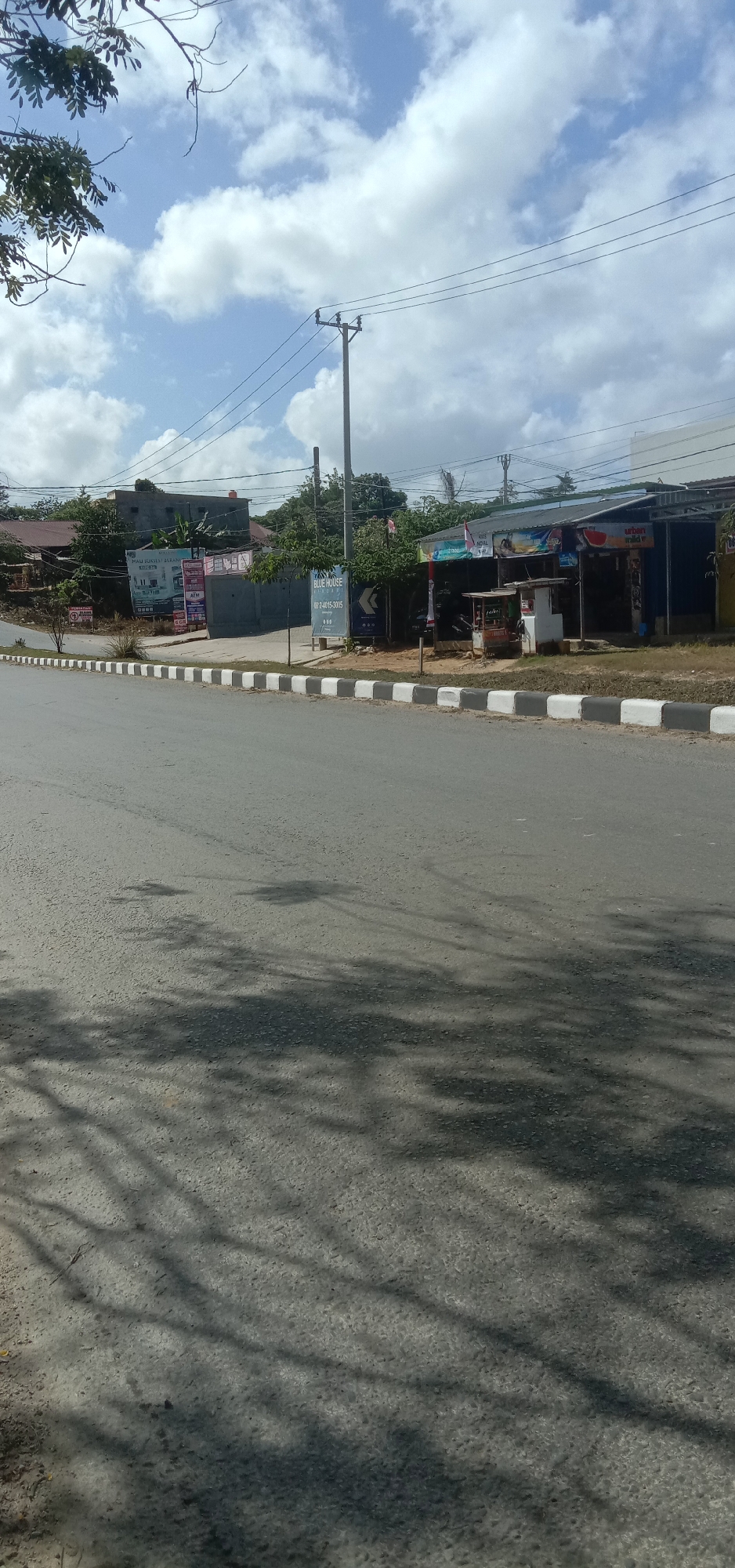 The image depicts a street scene in Kendari, Indonesia, on a partly cloudy day. The foreground is dominated by an asphalt road with distinct white and gray painted curbs. Shadows of trees and branches are cast across the road. In the mid-ground, a row of low-rise buildings line the side of the road, appearing to be shops or small businesses. These buildings have corrugated metal roofs and some have awnings. Signs are visible on the buildings, including one with the text "BLUE HOUSE" and a phone number, and another that appears to advertise housing. A utility pole with electrical wires stands near the buildings. A small food cart is visible in front of one of the shops. In the background, more buildings and dense vegetation can be seen, with trees forming a natural backdrop. The sky is a mix of blue and white clouds. There are no people or vehicles clearly visible in the frame.
