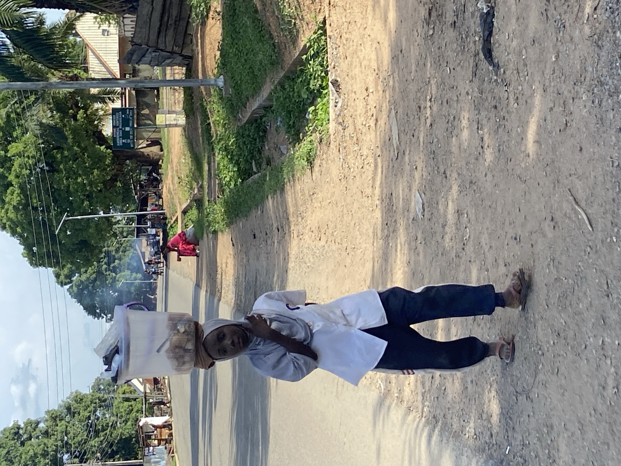 A woman is walking on the shoulder of a paved road in Mararaba, Nigeria, balancing a large, clear plastic container on her head, which appears to hold food. She wears a white top, dark pants, and sandals. A scarf covers her head. The road is flanked by greenery and buildings are visible in the background. Other people and activity are present further down the road. The surface of the road shoulder is dry and dusty. A black plastic bag lays on the road.