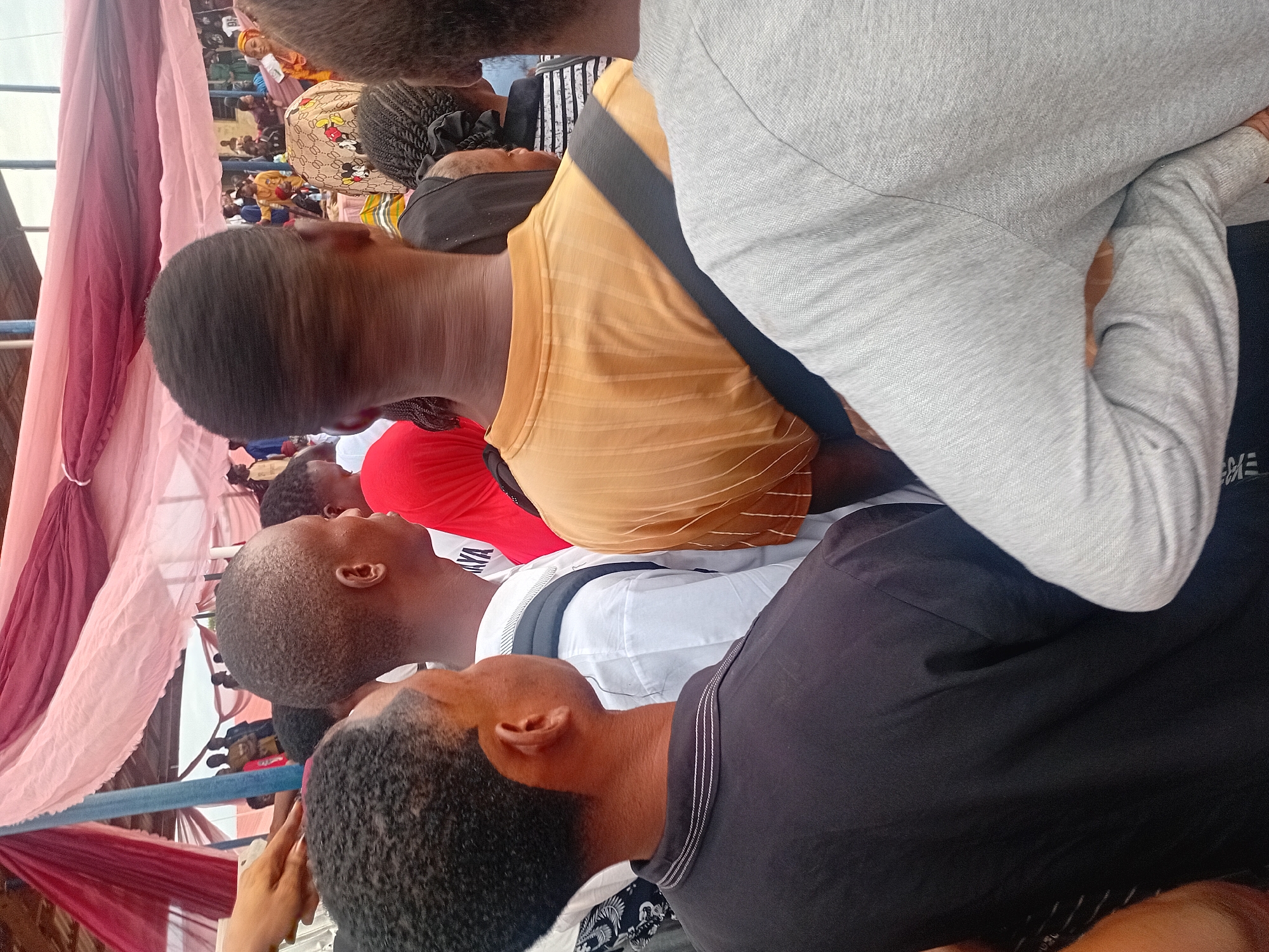 This image captures a crowded gathering in Ogbogbo, Nigeria, viewed from the perspective of an individual standing within the crowd. An overhead canopy, constructed from draped light pink and dark maroon fabric supported by blue metal poles, shelters the scene. In the foreground, multiple individuals are observed primarily from behind. A person on the far left wears a black short-sleeved polo shirt with white horizontal stripes on the collar, their head slightly turned to the right. Next to them, a person in a white long-sleeved shirt has a black strap, consistent with a backpack, over their right shoulder. To their right, a person in a mustard yellow ribbed t-shirt has a diagonal black strap across their back, extending from the left shoulder towards the right hip. Another individual, partially visible on the far right, wears a grey long-sleeved shirt with their left arm bent, hand resting on their hip. In the midground, additional attendees are visible, including one wearing a light-colored head covering patterned with Mickey Mouse figures. The background shows a dense crowd of people, some wearing blue, yellow, and green clothing, suggesting a communal event or assembly. Natural daylight illuminates the scene.