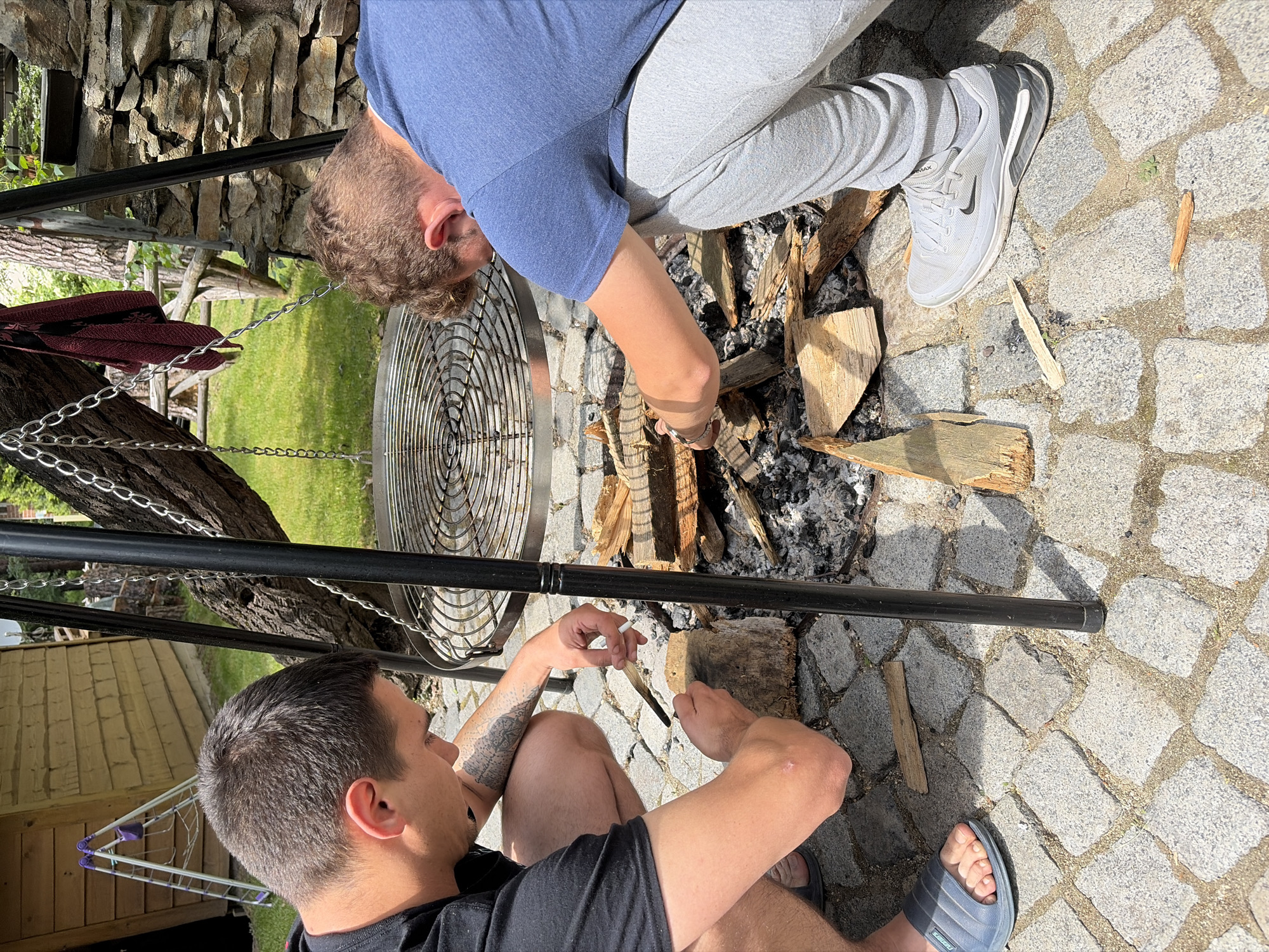 Two individuals are engaged in preparing a fire pit outdoors. One person, wearing a black t-shirt and gray shorts, is crouched on the left, holding a small piece of wood and a lighter. They are focused on the fire pit, which contains wood and ashes. The second person, in a blue t-shirt and gray sweatpants, is also crouched, on the right side of the frame, and is arranging pieces of wood in the fire pit. A metal grill is suspended above the fire pit by chains. The ground is paved with cobblestones. In the background, there is a wooden structure, a stone wall, and greenery, suggesting an outdoor setting.