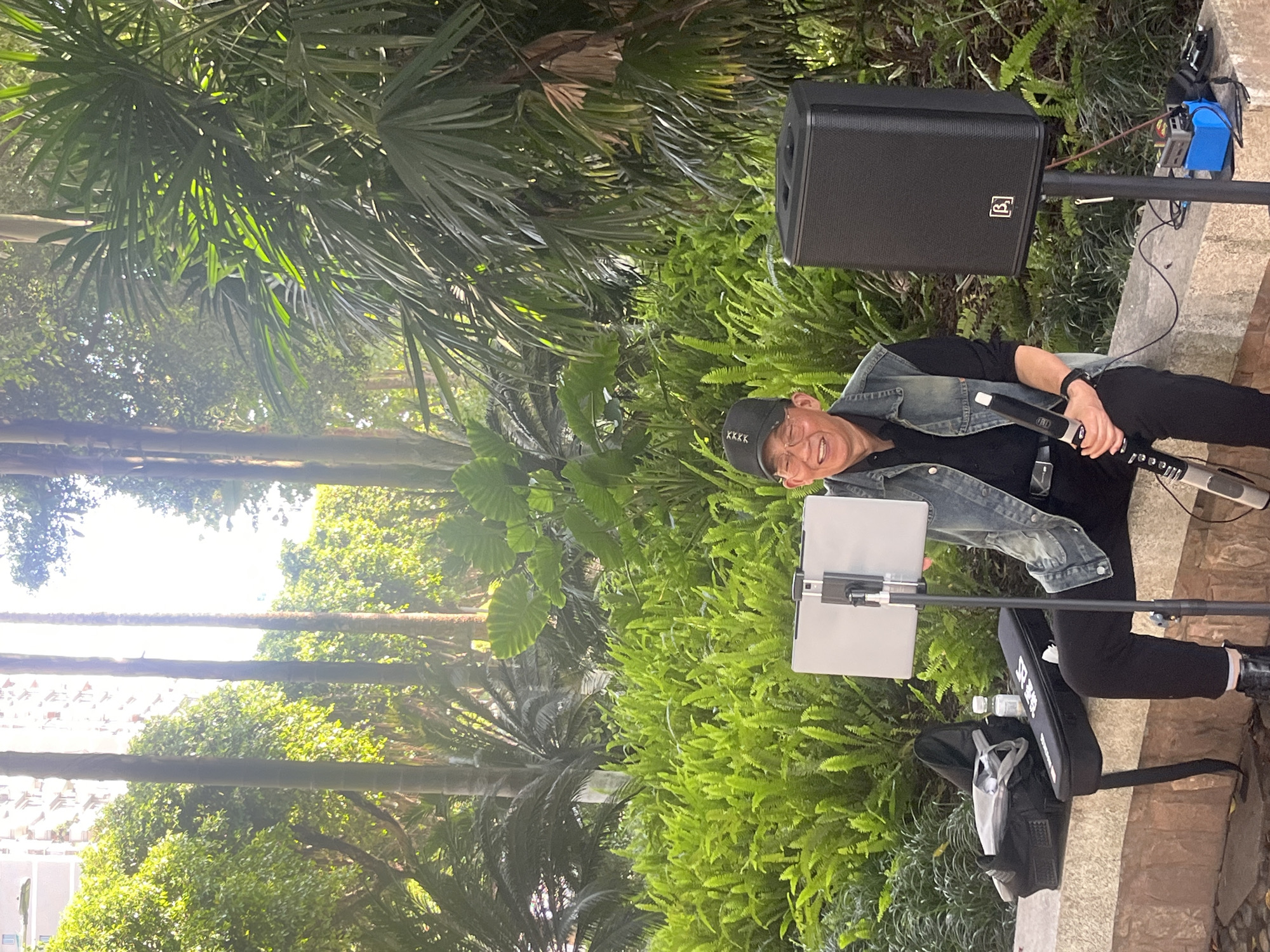 An adult man is depicted outdoors in what appears to be a lush park or garden setting in Jiangjin, China, during a bright daytime. He is smiling broadly, conveying a sense of joy or enthusiasm, as he appears to be performing or practicing music.
The man wears a black cap with "KKKK" text, glasses, a denim jacket over a black shirt, and black pants. He holds a black microphone in his right hand. To his right, a black portable speaker, featuring a logo resembling a stylized "B" or "JB," is mounted on a stand. Next to him, a digital tablet is secured on a separate stand, likely for displaying sheet music or lyrics. On the ground, a black carrying case, possibly for a musical instrument, is visible with "YAMAHA" printed on its side, and two clear water bottles rest on it. Additional blue equipment and cables are present near the speaker stand.
The background is abundant with various green plants, including dense ferns and palm trees, suggesting a subtropical or tropical environment. A stone or concrete path and low wall define the immediate foreground. Through the trees in the distance, glimpses of urban buildings can be seen under a bright, clear sky, indicating the park is situated within a city or town. The strong natural lighting suggests a clear day, likely in the morning or afternoon.
