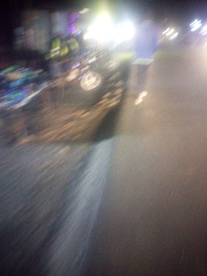 A blurry night scene depicts a vehicle with its headlights on. A barrier stands on the right side of the road. The asphalt pavement is visible, taking up the majority of the lower part of the frame. The scene has significant motion blur, and the overall lighting is poor. The colors are muted, mainly consisting of grey, black, and white. The image is difficult to interpret due to the blur.
