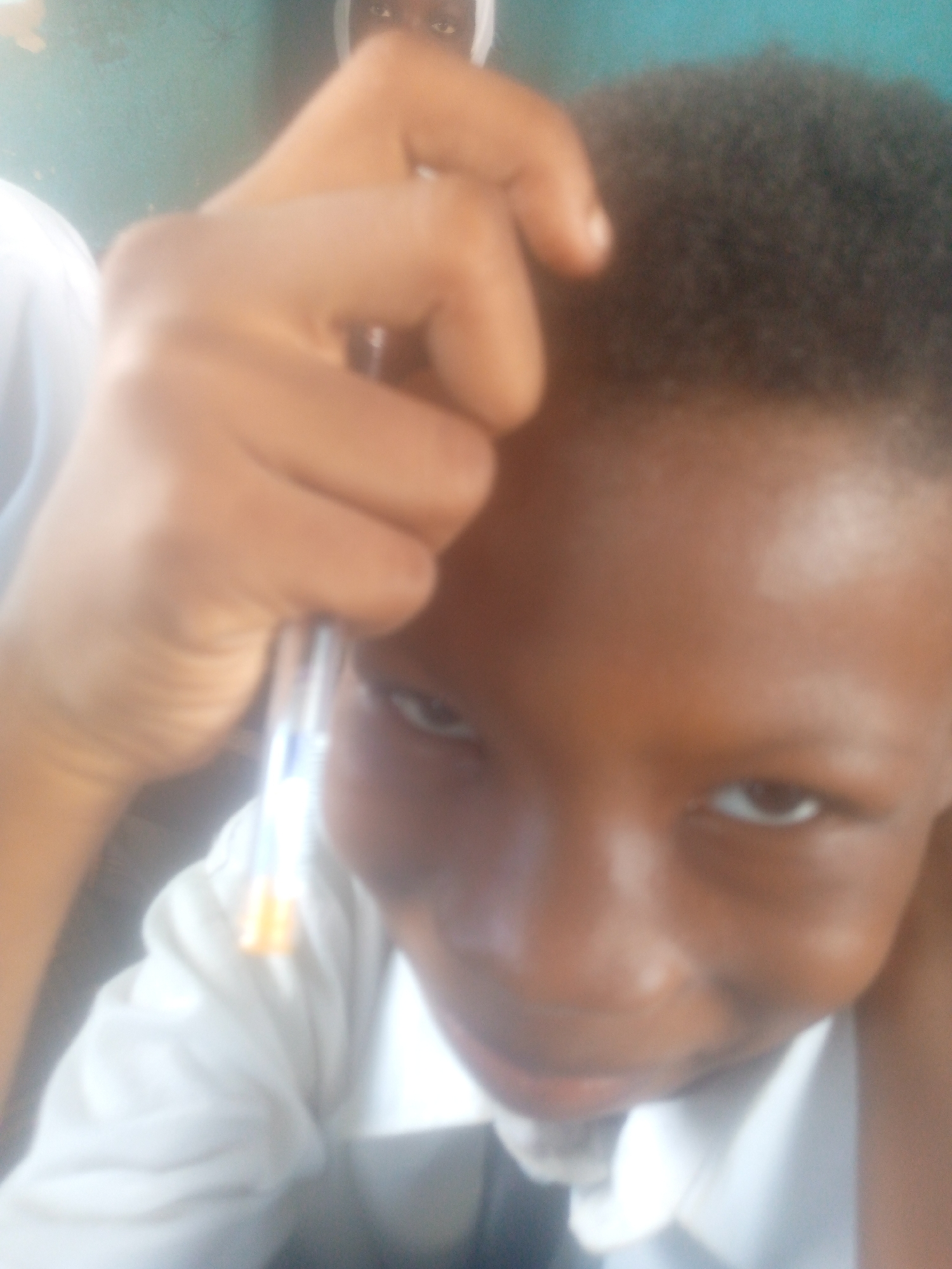 The image presents a close-up view of a child with short, dark hair, holding a pen near their face. The child is wearing a white collared shirt. The background is blurred, showing parts of another person with a white covering on their head. The location is unclear due to the lack of discernible details.