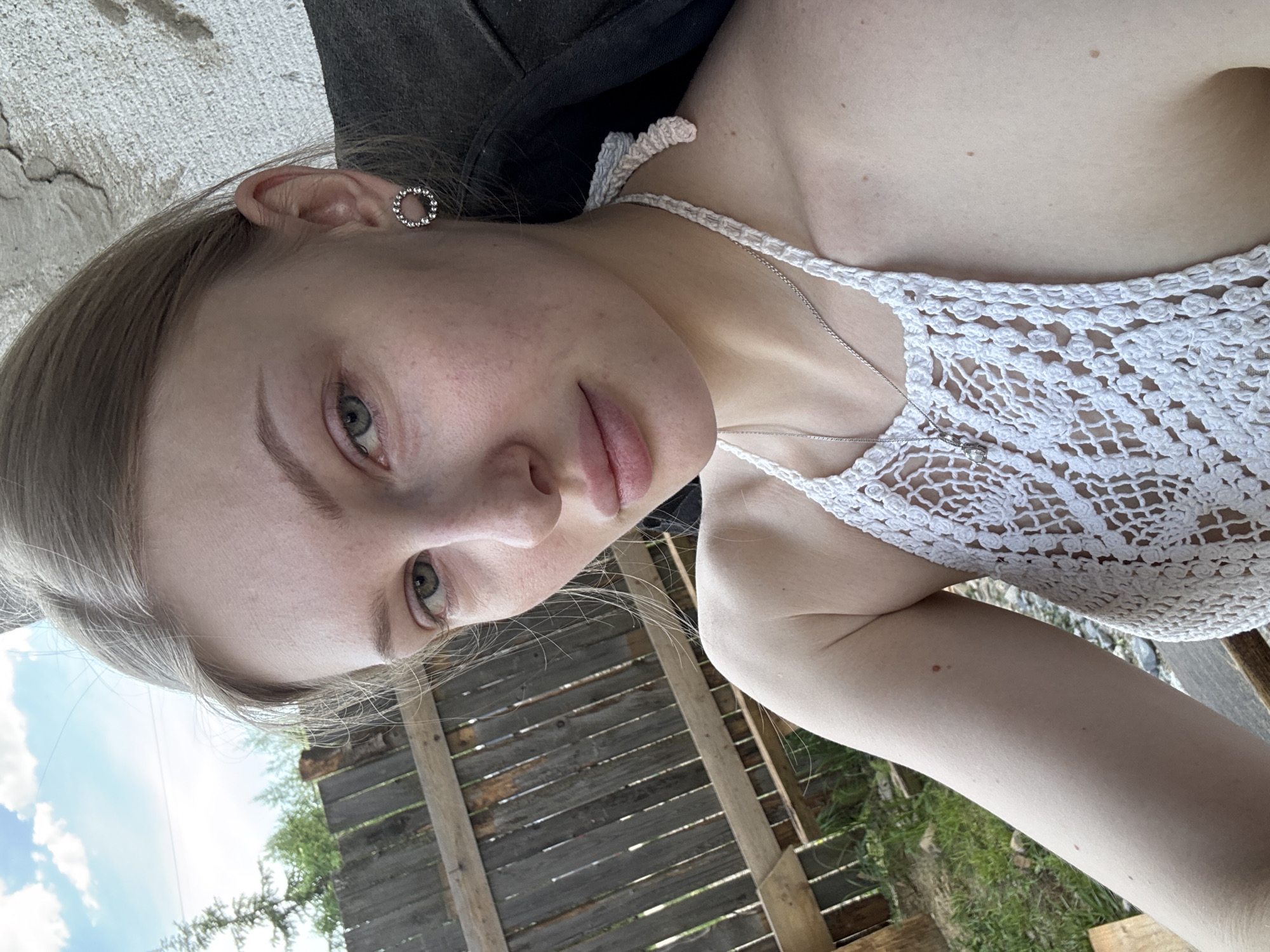 A medium, slightly angled shot shows a fair-skinned young woman outdoors. She has light brown or blonde hair. She is wearing a white, lace-like halter top and a silver necklace with a pendant. She has a circular earring in her left ear. The background includes a weathered wooden fence. Green vegetation is visible behind the fence. A patch of sky is seen in the upper left of the frame, implying the location is outdoors. Without any further clues, the location in the city of Perm, Russia cannot be determined.
