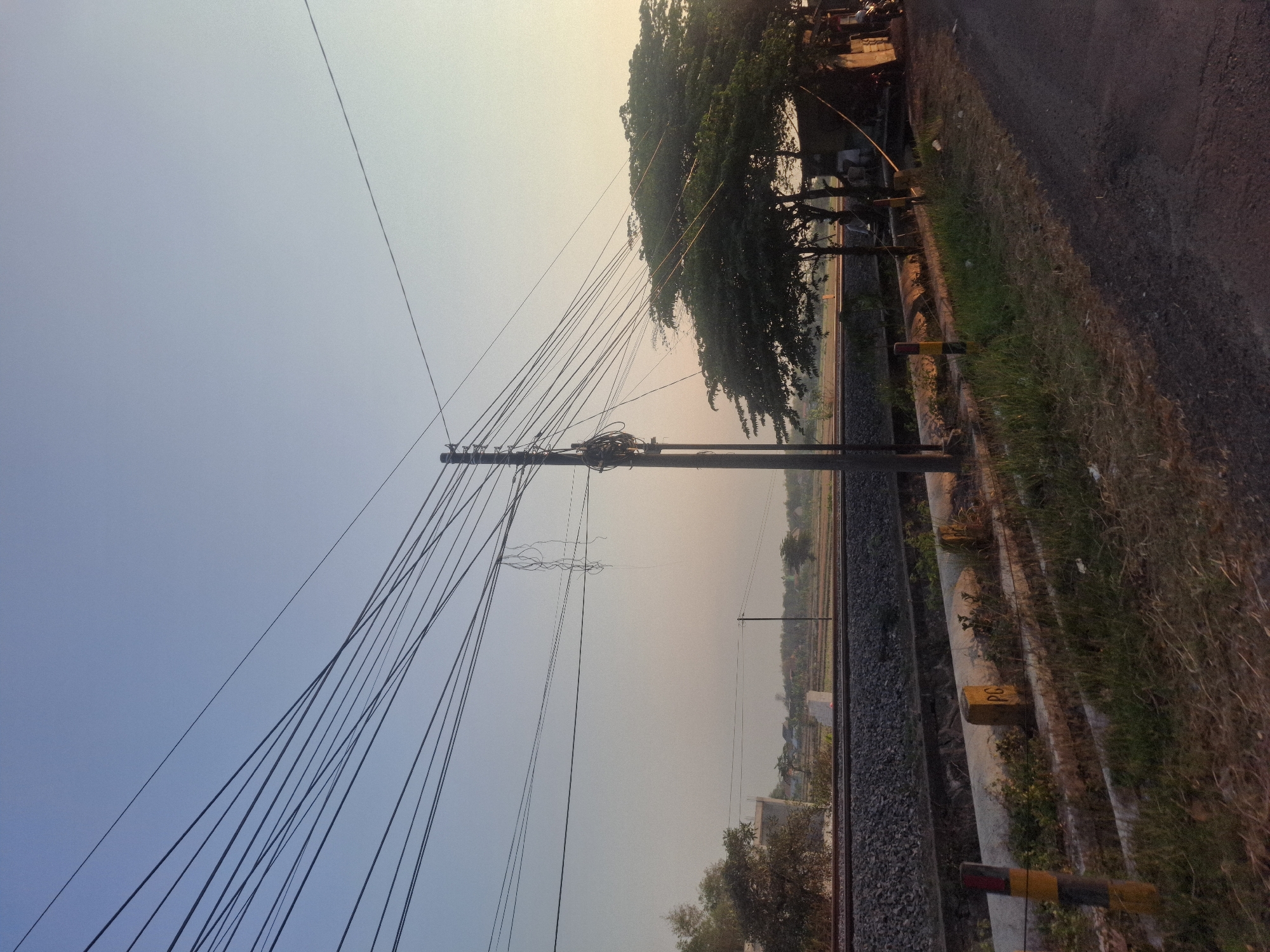 The image captures a serene, early morning scene in Waruwetan, Indonesia, likely near a rural railway line. The sky is a clear, pale blue, transitioning to a soft yellow hue near the horizon, suggesting the sun is either rising or setting. A prominent feature is a cluster of telephone or power lines crisscrossing the frame, strung from a tall utility pole. The pole itself is laden with bundles of wires and some loose, dangling cables.
In the mid-ground, a railway track with ballast stones runs horizontally across the image. Beyond the tracks, agricultural fields or open land are visible, with some scattered trees and low-lying vegetation. A faint, modern-looking structure, possibly an overpass or building under construction, is visible on the left side of the railway.
To the right of the utility pole, a lush green tree with dense foliage stands. Underneath its branches, a small, open-sided structure or shelter is partially visible, with at least two individuals appearing to be seated or standing nearby, though their features are not distinct. A motorcycle is parked to the far right of this structure.
In the foreground, a paved road or pathway runs diagonally from the bottom right corner, bordered by a grassy verge. Along the edge of the railway line, there are concrete barriers and posts, some marked with yellow and red stripes, and one with the letters "PG" clearly visible. The overall atmosphere is peaceful and rural, with the utility lines adding a touch of human infrastructure to the natural landscape.