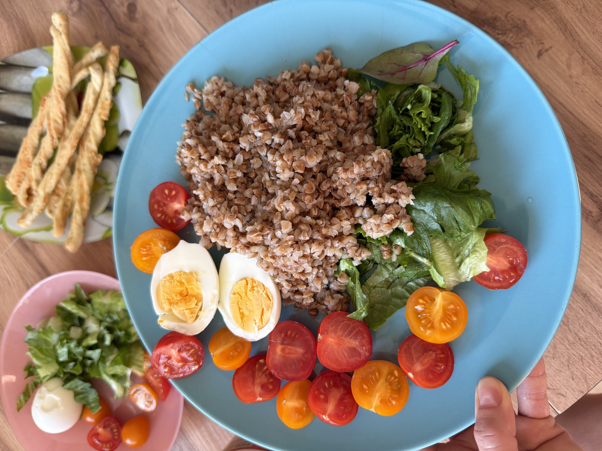 On a wooden surface, there are two plates of food and some breadsticks. One plate, a light blue color, contains buckwheat, lettuce, red and yellow cherry tomatoes, and hard-boiled eggs. A second, smaller pink plate contains lettuce, cherry tomatoes and a halved hard boiled egg. Breadsticks lie on a small white and green plate. The thumb and index finger of a fair-skinned hand are visible on the lower right edge of the blue plate. The setting suggests an indoor environment. There is no visual evidence to confirm that the location is Izhevsk, Russia.