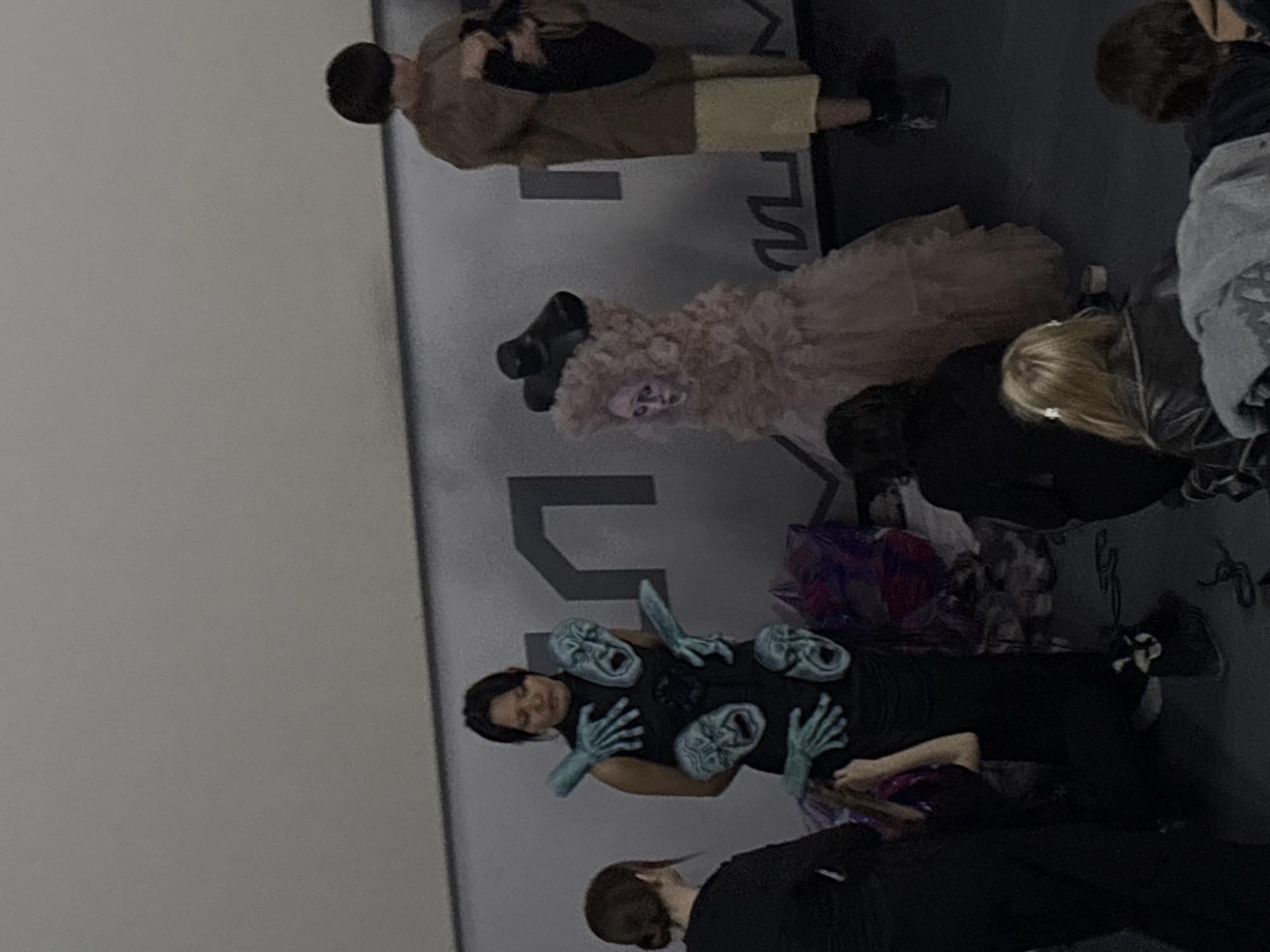 The image captures a scene in the city of Perm, Russia, featuring several individuals and elements suggesting an artistic or performance-related context. At the lower center, a person is dressed in a black outfit adorned with blue-gray masks and hand props, striking a pose. Above them, a figure in a voluminous, light pink dress with a prominent hat adds to the theatrical atmosphere. Other individuals are scattered around, seemingly observing or participating in the event. The backdrop includes a large, stylized letter "M" logo, possibly related to a brand or event. The overall setting appears to be indoors, possibly within an event space or art gallery.