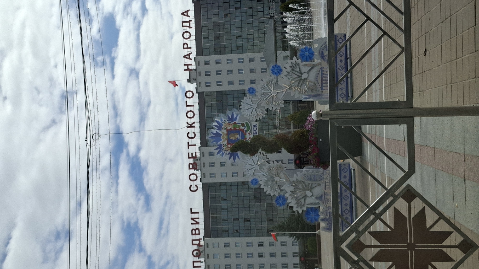 A wide shot captures the facade of a large, modern, light-colored building under a cloudy sky, with overhead wires crossing the frame. Red Cyrillic lettering across the top of the building reads "ПОДВИГ СОВЕТСКОГО НАРОДА" (feat of the Soviet people). In front of the building, a large, ornate archway is decorated with white and blue snowflake-like motifs, topped by a coat of arms featuring a lion, horse, and shield. A spiral-shaped tree stands in front of the coat of arms, with a planter of pink flowers at its base. To the right of the arch, a fountain sprays water upwards. A small red flag flies on a flagpole to the left of the arch, next to some evergreen trees. The foreground shows a paved area with a decorative metal railing featuring star-like patterns. The scene is likely Vitsebsk, Belarus, given the prominent inscription.