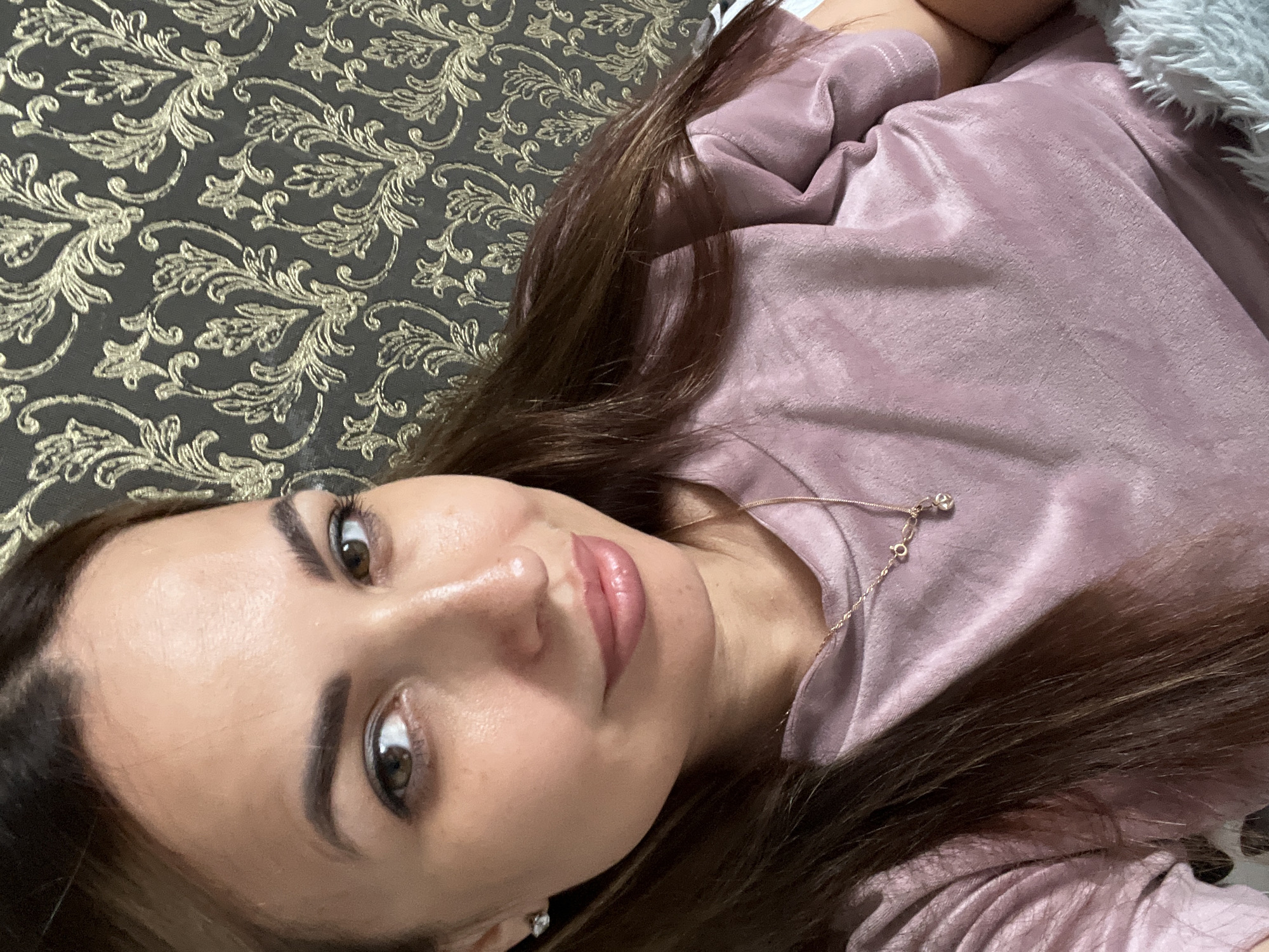 A medium shot captures a woman indoors, her face turned slightly towards the camera, set against a dark, ornate wallpaper with a gold damask pattern. She has long, dark brown hair that drapes over her left shoulder. Her skin appears smooth, and she is wearing subtle makeup. Her eyes, a warm brown, are looking directly at the viewer. A delicate rose gold necklace with a small pendant rests on her chest. She is wearing a soft, rose-colored, velvety shirt. To the right of the frame, a portion of a gray, fluffy blanket is visible. The lighting is soft, highlighting her facial features and creating a gentle sheen on her skin and clothing. The overall impression is one of relaxed intimacy.