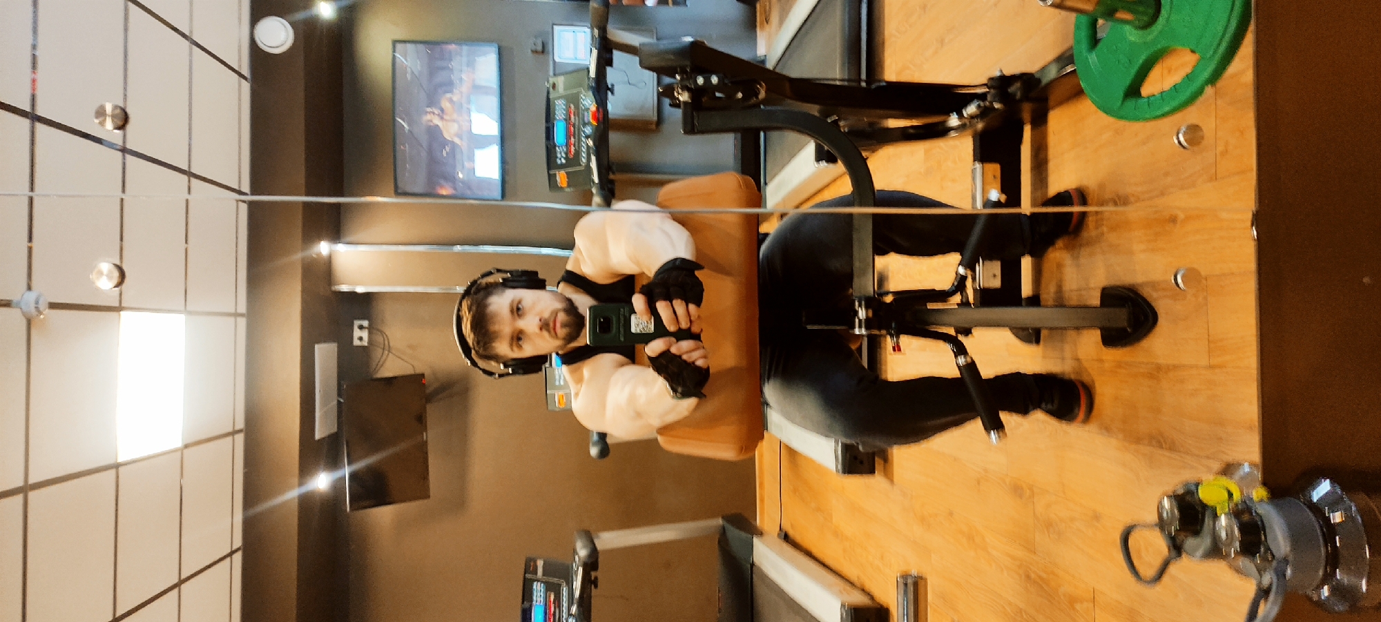 Here is a detailed description of the image:
**Setting:** The image appears to be taken inside a gym in Fryazino, Russia. The gym has a wood floor and partially mirrored walls, revealing a multi-paneled mirror with circular fasteners. Other walls are painted in dark neutral tones. Gym equipment is visible, as well as a television mounted on the wall.
**Subject:** A man wearing headphones and gym gloves is seated on a weight training machine. He is holding a cell phone, taking a selfie in the mirror. He is wearing a tank top that reveals his well-muscled arms.
**Objects:** Visible objects include: the gym equipment (a weight training machine with a green weight plate), a cell phone, headphones, gym gloves, a TV screen on the wall, mirrors, and light fixtures.
**Action:** The man is taking a selfie in the gym.
**Interaction:** The man interacts with his cell phone and the gym equipment.
**Environment:** The environment suggests a commercial gym setting with various machines and equipment for physical exercise.