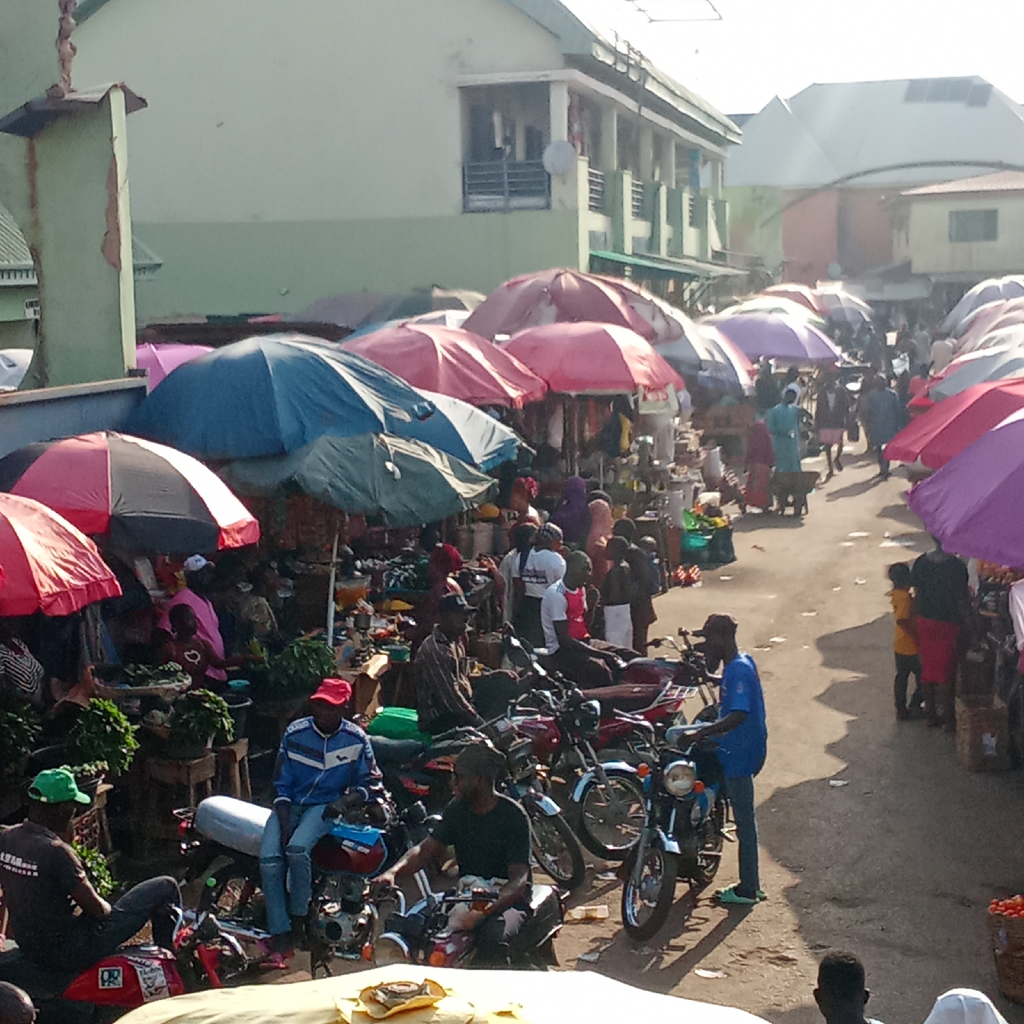 The image depicts a busy street market scene in Akwanga, Nigeria. Many vendors have erected colorful umbrellas for shade over their stalls. People are walking and interacting within the market, some carrying goods. Several motorcycles are parked in the foreground. The buildings in the background are two-story structures. The atmosphere appears crowded and active.