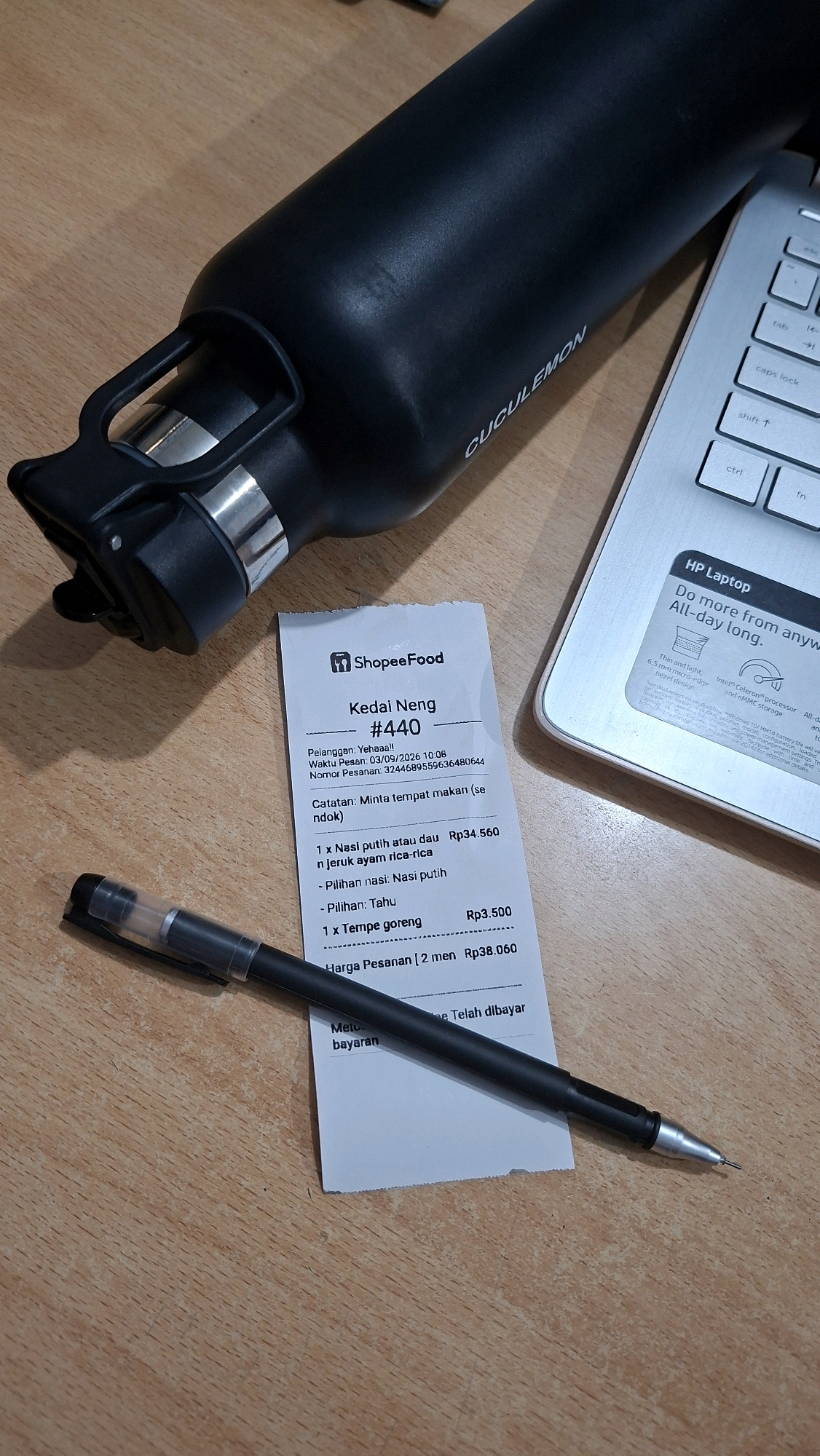 The image depicts a receipt from ShopeeFood, a food delivery service, on a wooden surface. The receipt is for an order from "Kedai Neng #440" and includes details about the ordered items: "1 x Nasi putih atau dau n jeruk ayam rica-rica" costing Rp34.560, with a note that the rice choice is "Nasi putih" and the tofu choice is "Tahu". Also listed is "1 x Tempe goreng" for Rp3.500. The total cost of the order for "2 men" is Rp38.060. Below this, it says "Metu... E Telah dibayar", indicating payment has been made.
In addition to the receipt, a black pen is lying on top of it, with its tip pointing towards the bottom right of the image. A black water bottle with a silver band and a black handle is visible in the upper left corner of the frame. Part of a silver laptop keyboard, with keys like "fn", "ctrl", "shift", "caps lock", "tab", and "esc" visible, is in the upper right portion of the image. The overall environment appears to be a desk or table, suggesting an indoor setting. The presence of a ShopeeFood receipt strongly suggests the scene is located in Indonesia, and given the context of food delivery services, it could be in an urban or suburban area. The provided location in Karet, Indonesia, further specifies this.