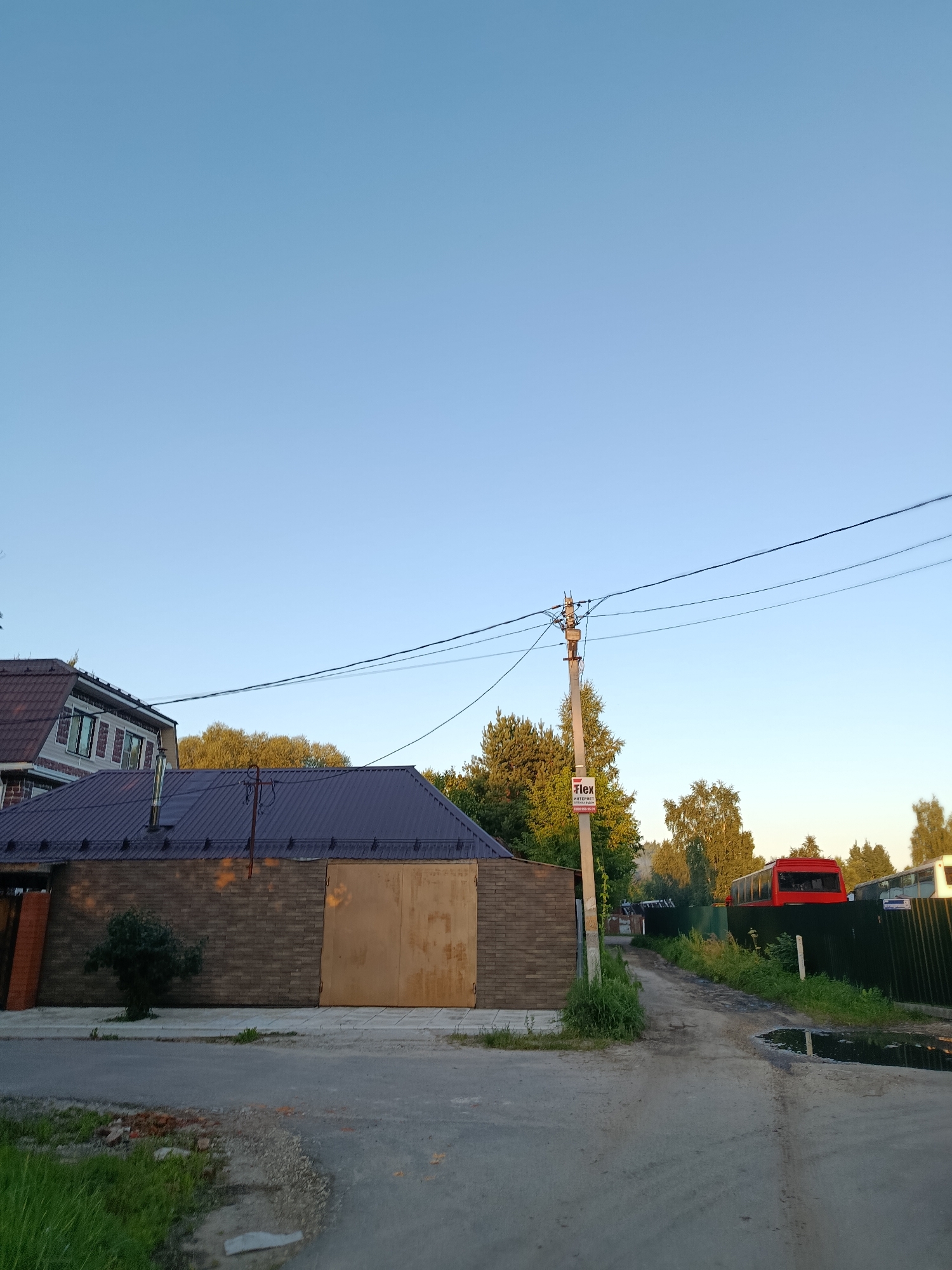The image shows a street scene in the city of Vereya, Russia. In the foreground, there is a paved road with a puddle on the right side. To the left of the road, there is a patch of grass and some scattered bricks. In the middle ground, there is a building with a dark brown metal roof and a large beige metal garage door. A small bush is planted in front of the building. Further back, a power pole with electrical wires is visible, with a sign attached to it that reads "Flex Internet". Beyond the building, trees and a green metal fence are present. A red bus is visible behind the fence. The sky is clear and blue.