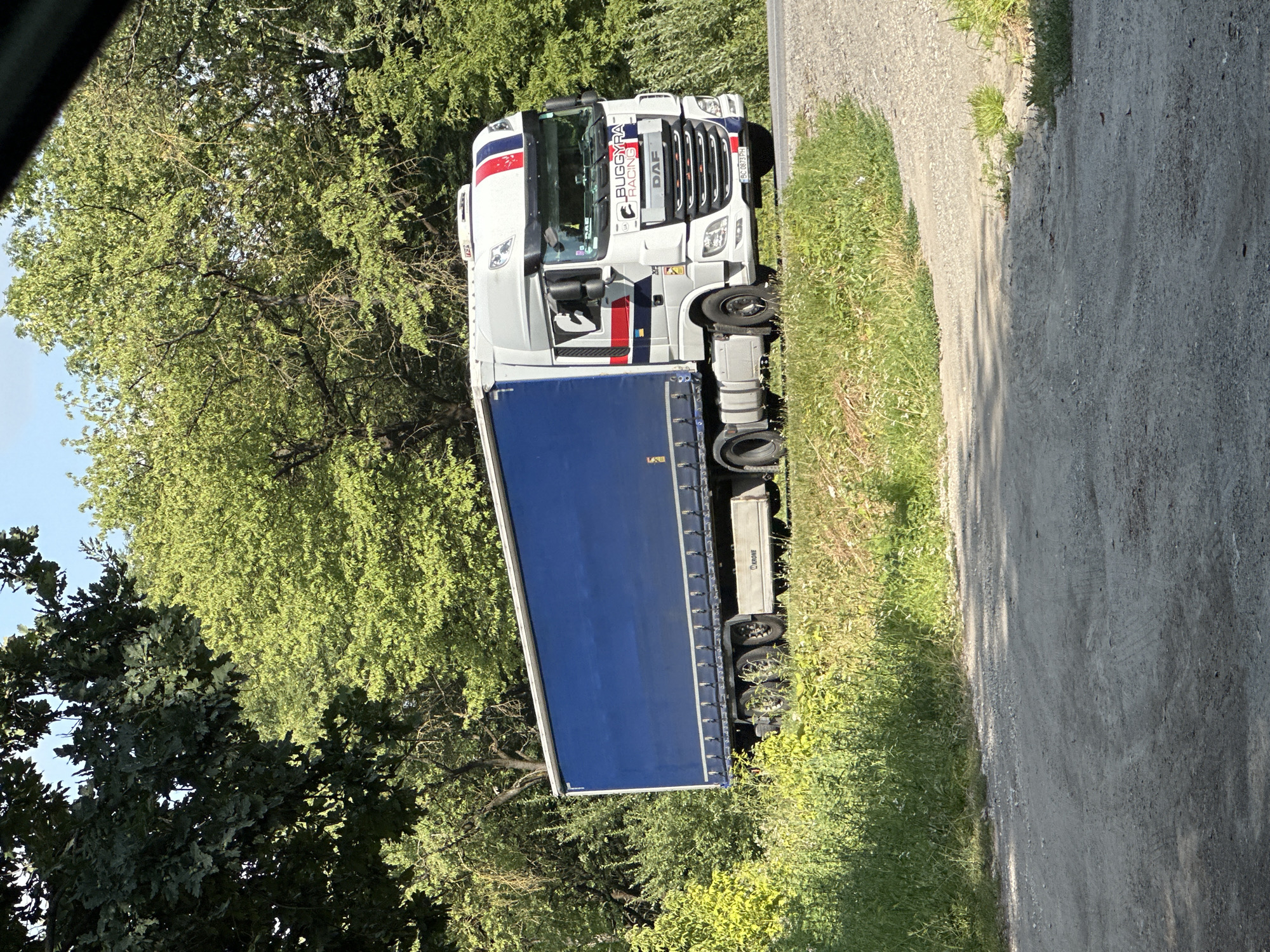A white DAF truck with a blue trailer is driving on a road. The truck has "BUGGYRA RACING" written on the side. The trailer is mostly blue and appears to be a curtain-sided trailer. The truck and trailer are in motion, as indicated by the slight motion blur. The truck has a license plate that reads "BC 0873 PH". The scene appears to be a rural or semi-rural area, with trees and vegetation on either side of the road. The road is paved with asphalt and gravel. The sky is blue and clear. This image was taken in Shatava, Ukraine.