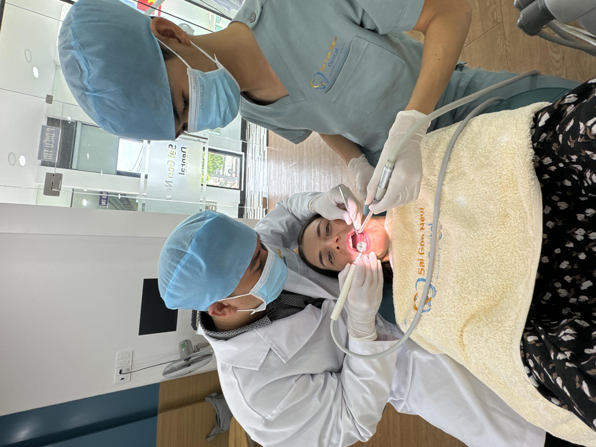 In a dental clinic in Phu Thuy Ward, Vietnam, two dental professionals are performing a procedure on a female patient. The image is oriented vertically, showing the patient lying horizontally with her head towards the bottom of the frame, and the professionals positioned above her.
The patient, a young woman with dark hair and light skin, is wearing a dark top with a white floral pattern. Her mouth is open, and she appears calm and cooperative. A yellowish-beige textured towel or blanket, inscribed with "Sai Gon New Dental," covers her chest.
One dental professional, wearing a light blue scrub top with a "Sai Gon New Dental" logo, a blue disposable hairnet, and a light blue surgical mask, is standing. They are wearing white disposable gloves and are holding a dental handpiece or suction device, directing it towards the patient's mouth. The other professional, wearing a white lab coat over a grey layer, a blue disposable hairnet, and a light blue surgical mask that reads "Hello Mask," is positioned near the patient's head. This professional is also wearing white disposable gloves and is using a dental mirror and another tool inside the patient's mouth.
The setting is a clean, modern dental office. The background features glass partitions and doors, reflecting light and parts of the clinic, including the mirrored text "Sai Gon New Dental" and other Vietnamese characters like "IAT U310.9". The walls are white with a blue accent strip along the bottom, and a small white electric fan is visible on the wall. The flooring is a light-colored wood. The overall scene depicts a professional dental examination or treatment during daytime hours.