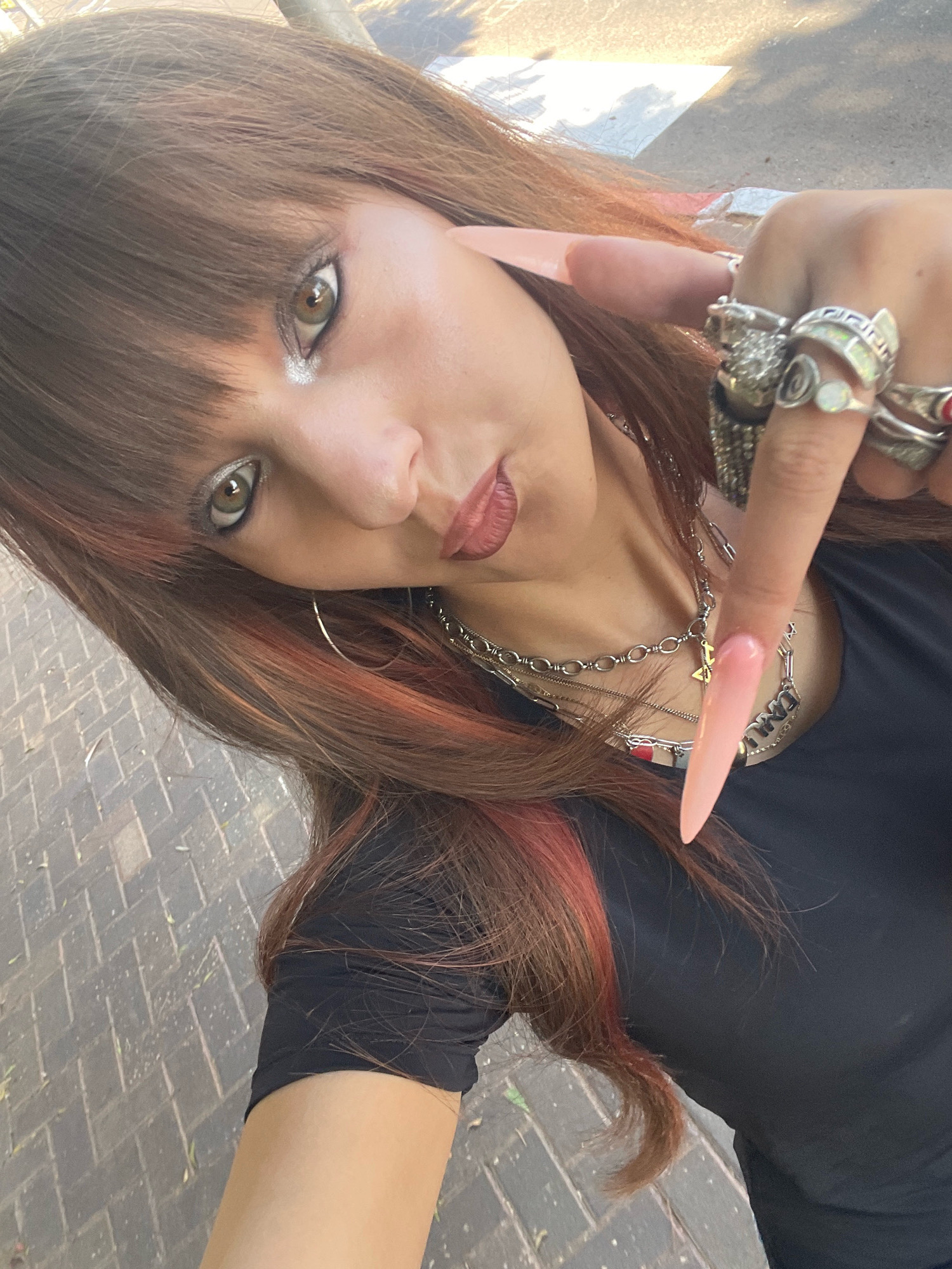 The image shows a close-up, angled selfie of a young woman with brown hair with reddish streaks, wearing a black t-shirt and multiple rings on her right hand. She is also wearing layered necklaces. Her left index finger is extended towards the camera, and her right hand is positioned near her face, adorned with rings. She has prominent eye makeup, including glittery eyeshadow. The background appears to be an outdoor setting with a paved walkway made of brick-like tiles, suggesting a public space such as a sidewalk or park path. The lighting indicates it is daytime. The presence of a tiled pathway is consistent with many areas in the city of Raanana, Israel, particularly along its sidewalks and public squares.