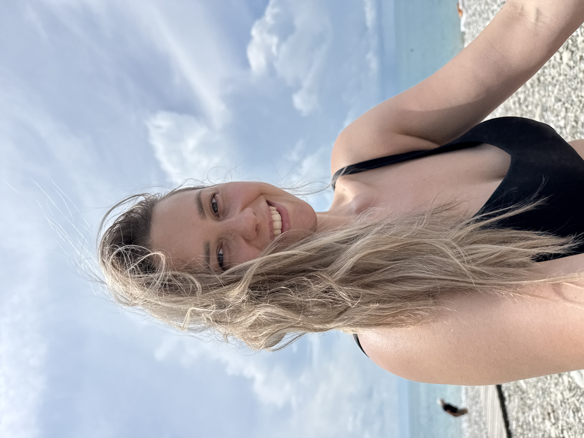 A woman with blonde hair smiles at the camera on a beach in Nebug, Russia. She is wearing a black swimsuit. The beach is covered in pebbles, and the sea is visible in the background. The sky is cloudy. In the background, another person is visible walking on a wooden boardwalk.