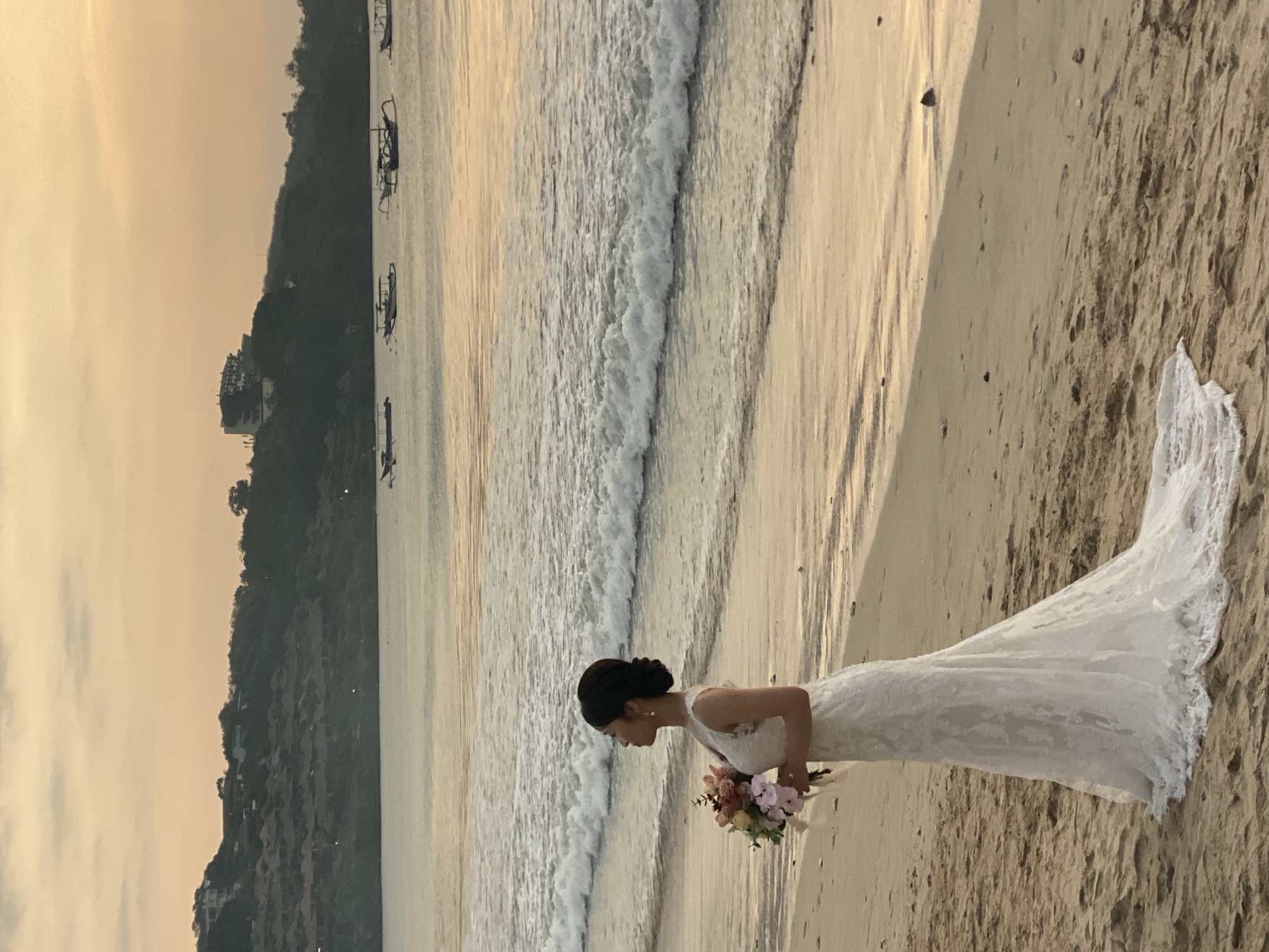 A full shot captures a bride on a sandy beach in Kuta Selatan, Indonesia, holding a bouquet of flowers. She is wearing a sleeveless, floor-length white lace wedding gown with a train. The bride is facing left, looking towards the ocean, with her hair in a bun. The ocean has small waves lapping at the shore, and several traditional Indonesian outrigger boats are visible in the distance. The background features a lush, green hillside dotted with buildings and a hotel on the summit. The sky is a soft, warm orange, indicating either sunrise or sunset. The overall lighting suggests it is either early morning or late evening.