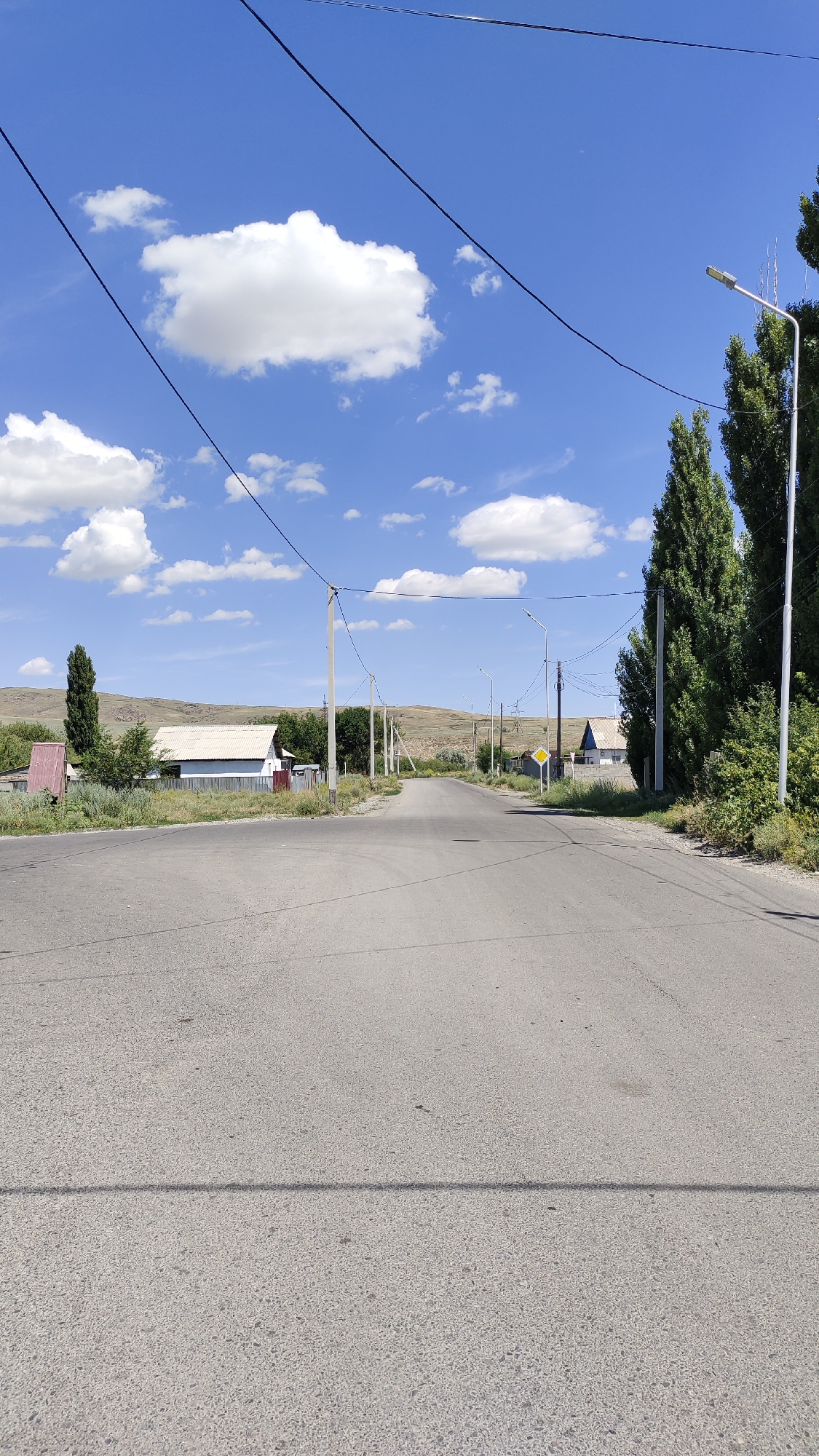 The image shows a view of a rural road in Taldykorgan, Kazakhstan, on a clear, sunny day with scattered white clouds. The road is asphalt and appears to be well-maintained. There are power lines running overhead, supported by utility poles. On either side of the road, there are houses, trees, and vegetation. In the distance, there are rolling hills. A yellow diamond-shaped sign is visible on the right side of the road, indicating a hazard or warning. There are no people or vehicles visible in the scene. The overall impression is one of a quiet, rural setting.