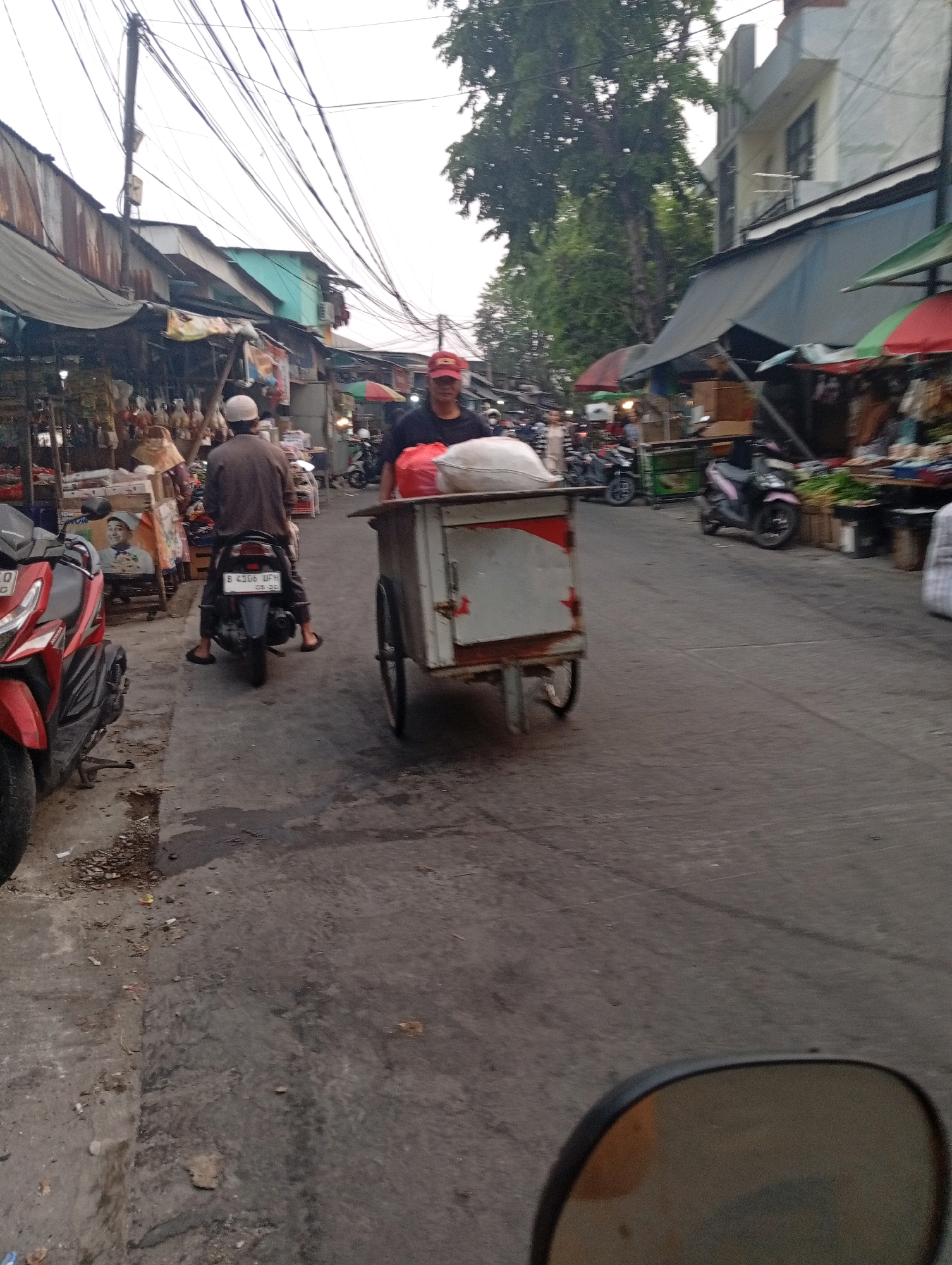 The image depicts a street scene in North Jakarta, Indonesia, likely near a market or roadside stalls. In the foreground, a red and black scooter is parked on the left. A person wearing a white cap and brown clothing is seated on a scooter in the middle ground, facing away from the camera. To the right of this person, a cart with a white body and red trim is being pushed by an individual wearing a red cap. The cart contains what appears to be a large white bag. The street is paved and shows signs of wear and tear. Along the sides of the street are various market stalls with awnings, displaying goods. Overhead, a complex network of electrical wires crisscrosses the sky, typical of urban environments in Indonesia. In the background, trees and buildings are visible. The overall impression is of a busy, functional street scene in an Indonesian city.