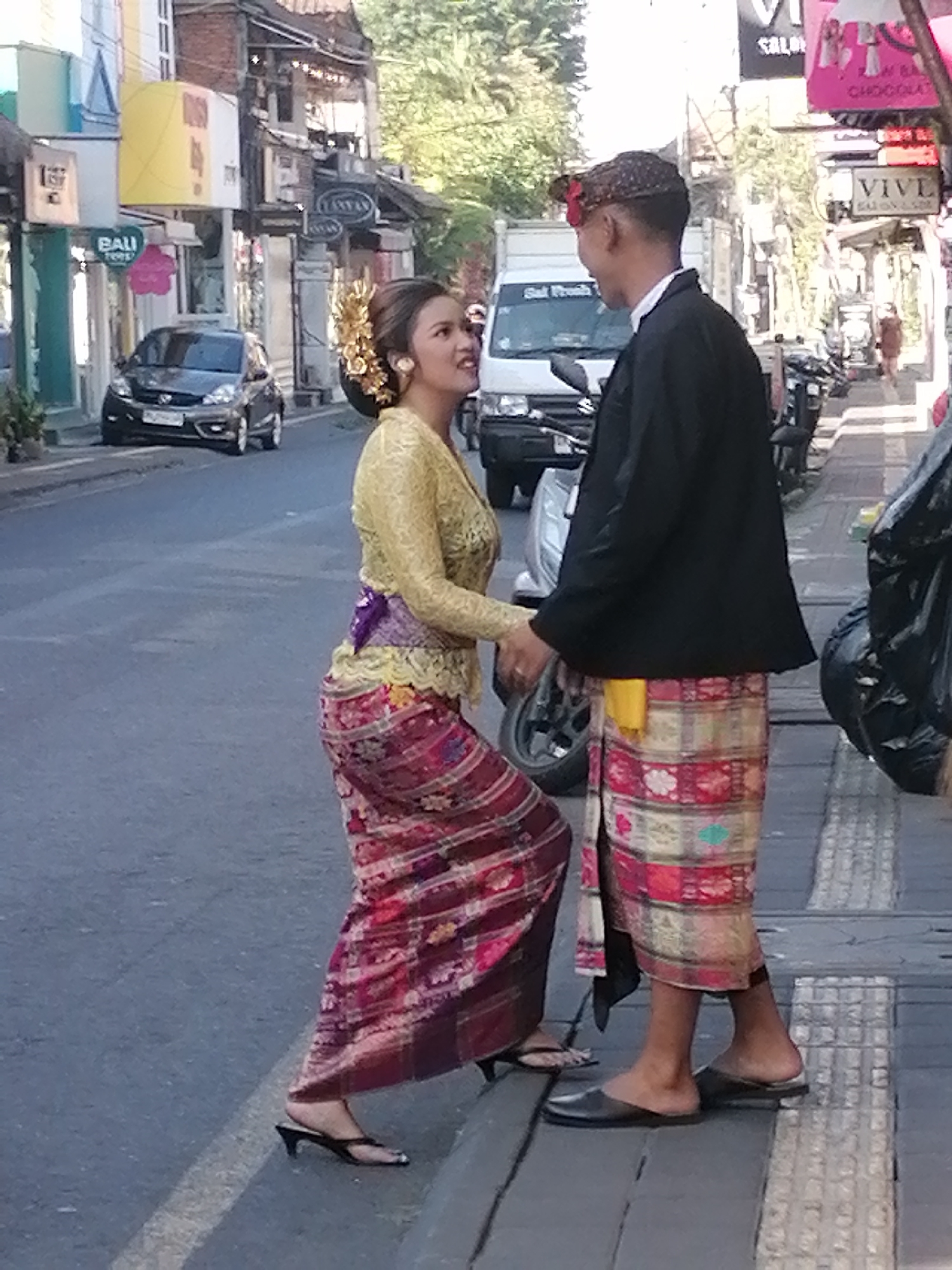 In the city of Ubud, Indonesia, a young woman and a man, dressed in traditional Balinese attire, stand facing each other on a street. The woman, with a gold headdress and earrings, wears a yellow lace top and a red, purple, and gold patterned sarong. She is looking towards her right, with a smile on her face. The man, wearing a black jacket and a patterned sarong, is looking towards the woman. They are standing near the sidewalk, with a street and buildings in the background. A car and a truck are visible in the background. The scene appears to be a street in a bustling market area.