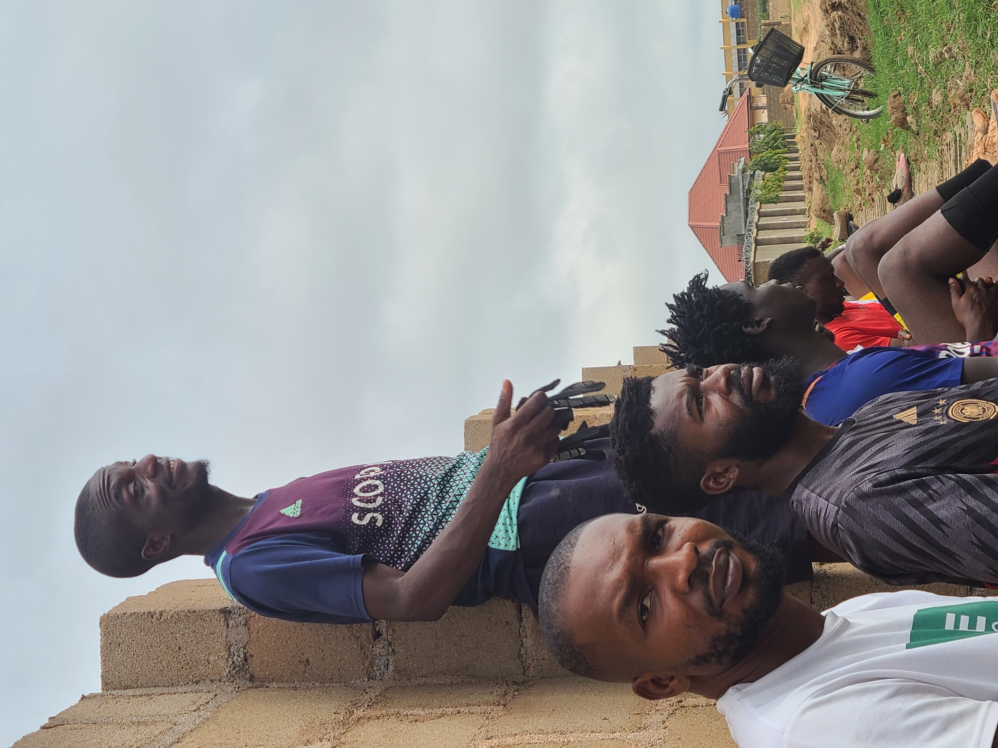 A group of men is gathered outdoors in what appears to be a semi-urban or rural setting. The image is taken from a low angle, looking up at the subjects and the sky.
In the immediate foreground, a man with a short beard is looking directly at the camera with a relaxed expression, wearing a white t-shirt with a green graphic or text on the left side (partially visible). Behind him and slightly to the right, another man with dreadlocks and a beard is visible, wearing a black and grey striped sports jersey with an Adidas logo and several gold stars. He is looking upwards and to the right. Further behind him, another man with dreadlocks is wearing a blue and red jersey, also looking in a similar direction.
On the far left, a man with short hair and a beard is leaning against or standing on a low brick wall, looking upwards with a smile. He is wearing a maroon and blue patterned t-shirt with "SCODS" and an Adidas logo visible on it. He appears to be holding a dark object, possibly a phone, in his right hand. Other partial figures are visible further back in the group, seemingly seated on the ground or a lower structure.
The background shows a light grey, overcast sky. To the right, there's a dirt mound with some grass, and a bicycle with a basket is parked on top of it. Beyond the dirt mound, several buildings with reddish-brown roofs are visible, suggesting houses or small structures. The men appear to be relaxing or conversing in an informal outdoor gathering. The setting includes construction materials like brick walls in the foreground.