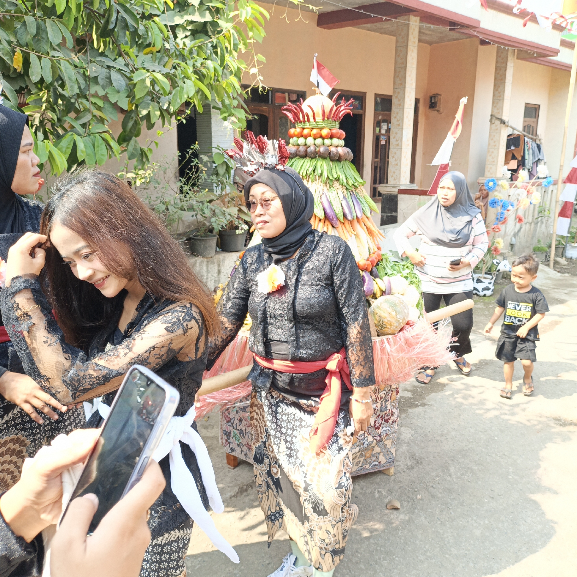 In the city of Tanahbaya, Indonesia, a festive procession is underway, likely celebrating a harvest or cultural event. The foreground features a young woman in a black lace outfit with intricate patterns and a red sash, her hand raised to her long, dark hair as she smiles downwards. Beside her, another woman in a similar black lace outfit is focused on her phone.
Behind them, a large, elaborately decorated float is being pulled, shaped like a tiered cone and adorned with a vibrant assortment of fruits and vegetables, including tomatoes, cucumbers, chilies, and eggplants. A small Indonesian flag flies from the top of the float. A woman in a grey hijab and patterned shirt is seen managing the float, holding a mobile phone. A young boy in a black t-shirt and shorts walks alongside the procession.
The background reveals traditional Indonesian houses with beige walls and tiled roofs, suggesting a residential area. Lush green trees and potted plants add to the outdoor setting. Indonesian flags and decorative elements are visible along the street, indicating a celebratory atmosphere. The ground is paved with concrete, and shadows from the structures and foliage cast across the scene.