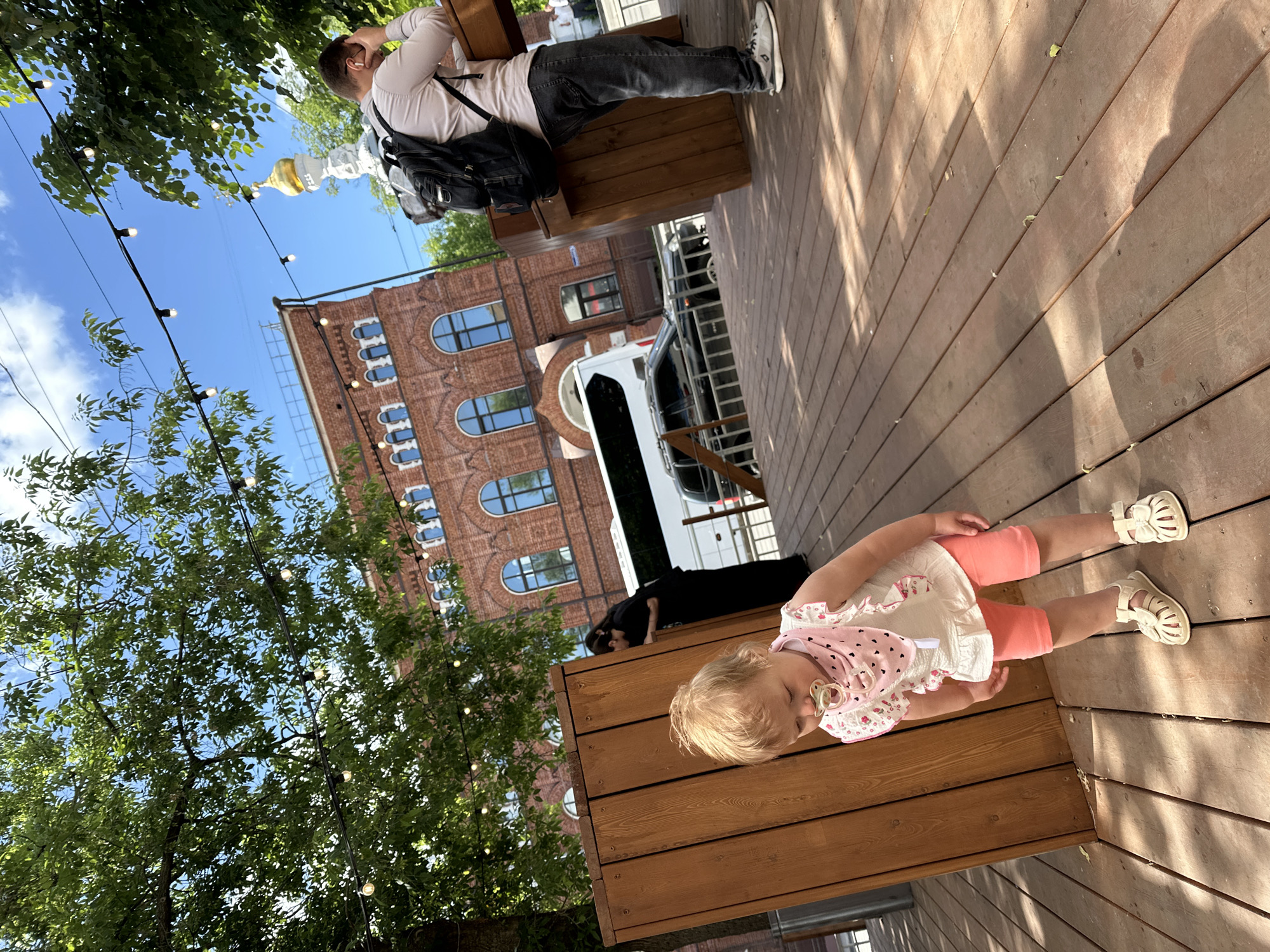The image captures an outdoor scene, likely a cafe or restaurant area, with a wooden deck floor and wooden benches. The bright daylight and shadows indicate a sunny day, likely mid-morning to late afternoon, under a clear blue sky with some scattered clouds. The setting is an urban environment, possibly in Nizhny Novgorod, Russia, given the architectural style of the brick building and the presence of a church dome in the distance.
In the foreground, a young child with blonde hair, wearing a white top with pink polka dots, pink shorts, and white sandals, stands next to a wooden bench. A pacifier is visible in the child's mouth, and they appear to be looking down.
In the middle ground, a man is seated on a wooden bench, facing away from the camera. He wears a light-colored shirt, dark jeans, and white sneakers, with a black backpack resting beside him on the bench. He is looking up or to the side, with his hand near his head. Further back, another person, dressed in black, is partially visible near a white bus or van parked on the street.
The background features a large, ornate brick building with multiple arched windows and intricate detailing. Lush green trees line the area, with string lights suspended between them, suggesting an evening ambiance option for the space. In the far distance, a golden church dome can be seen against the sky. No visible text is clearly discernible in the image. The overall atmosphere is relaxed and casual.